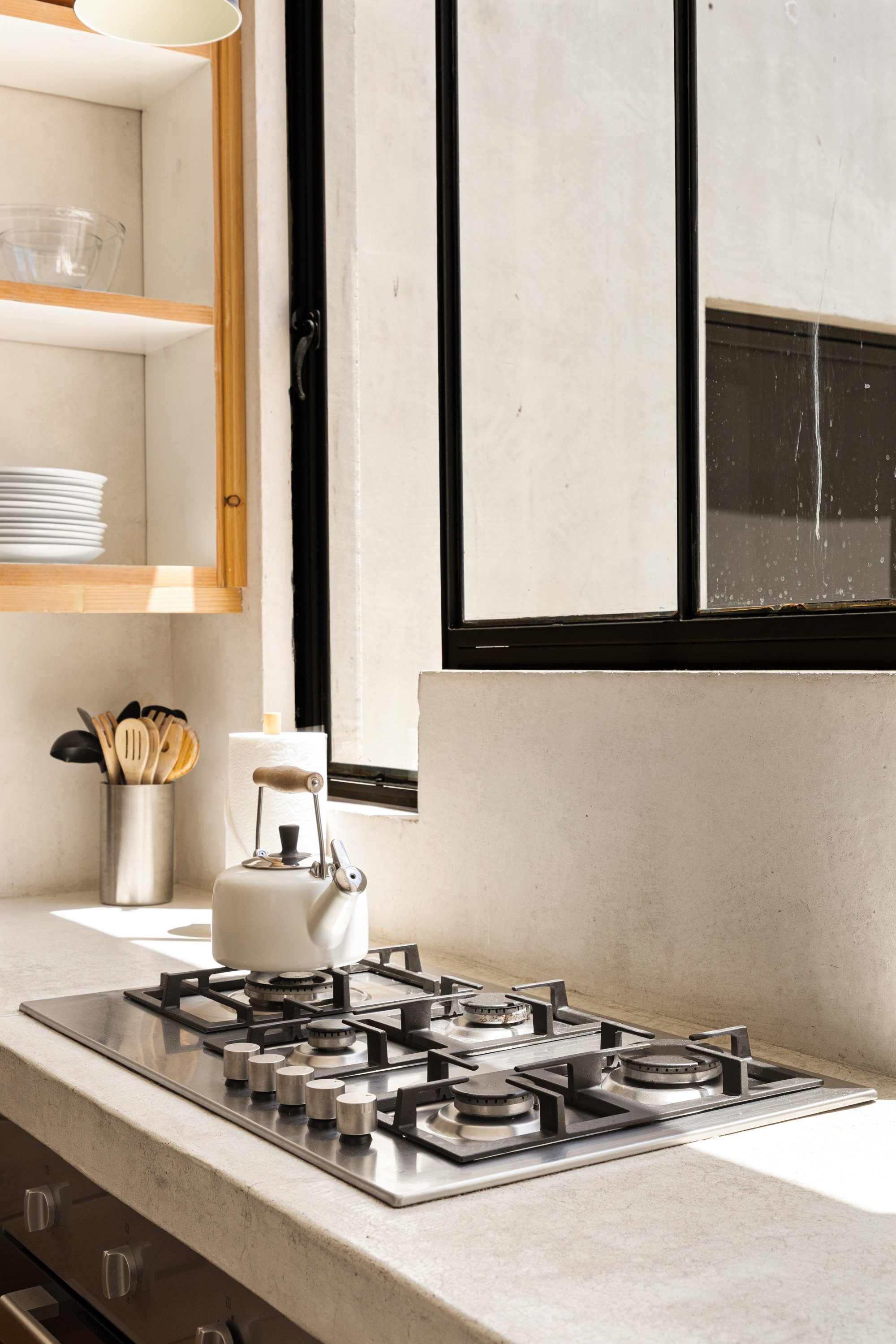 A kitchen counter with a white kettle on a gas stove, a soap dispenser, a paper towel holder, and a container with wooden utensils. There is a window with black frames to the right.
