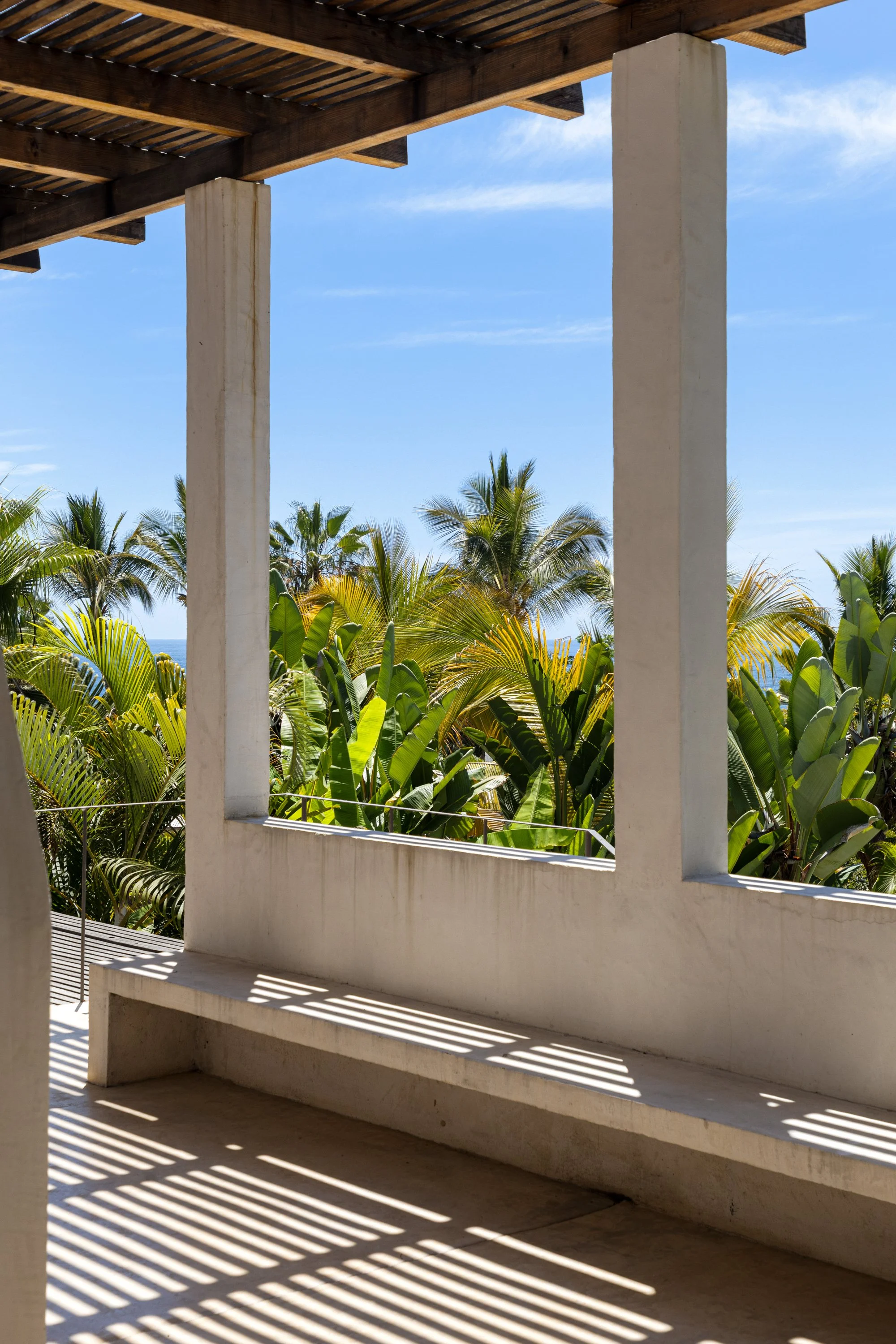 View from a balcony or porch with a concrete railing and wooden beams overhead, overlooking lush tropical plants and palm trees under a bright blue sky.
