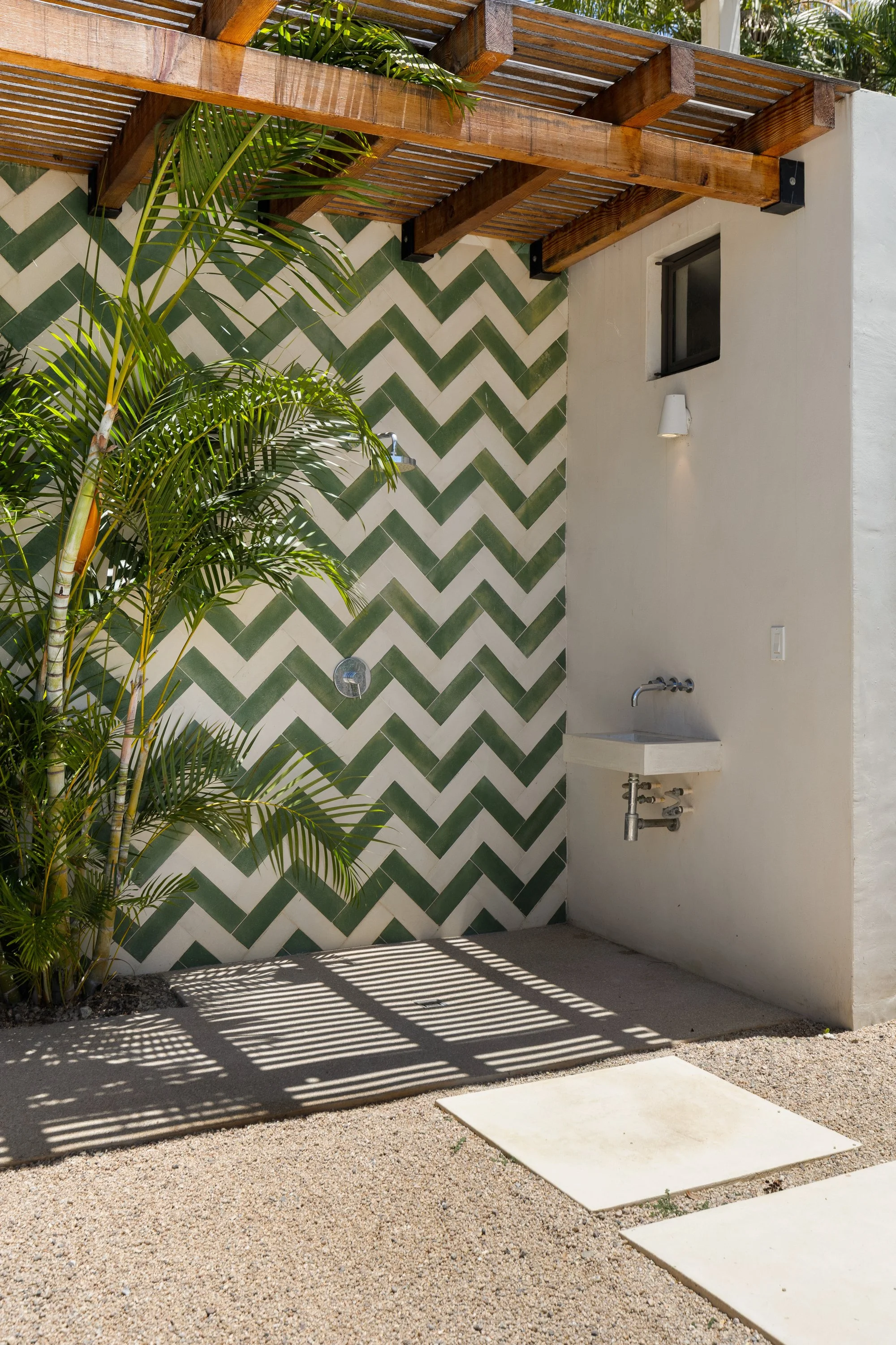 An outdoor bathroom area featuring a green and white zigzag tiled wall, a small white sink, a white wall-mounted light, a window, and tall green plants casting striped shadows on the gravel ground.