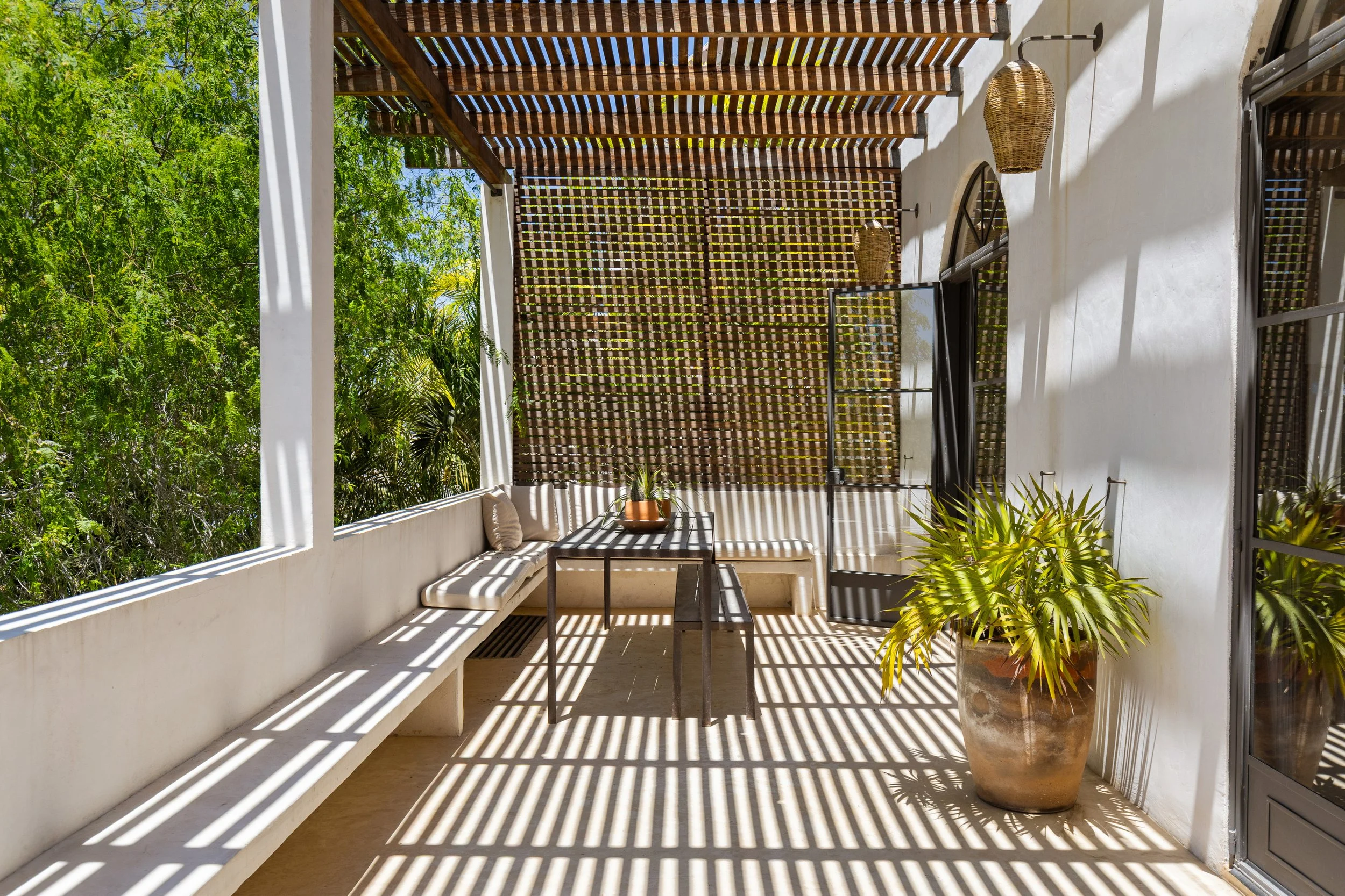 Balcony with white walls and wooden slatted ceiling creating patterned shadows; potted plant and bench with cushions; glass door revealing an outdoor green landscape.