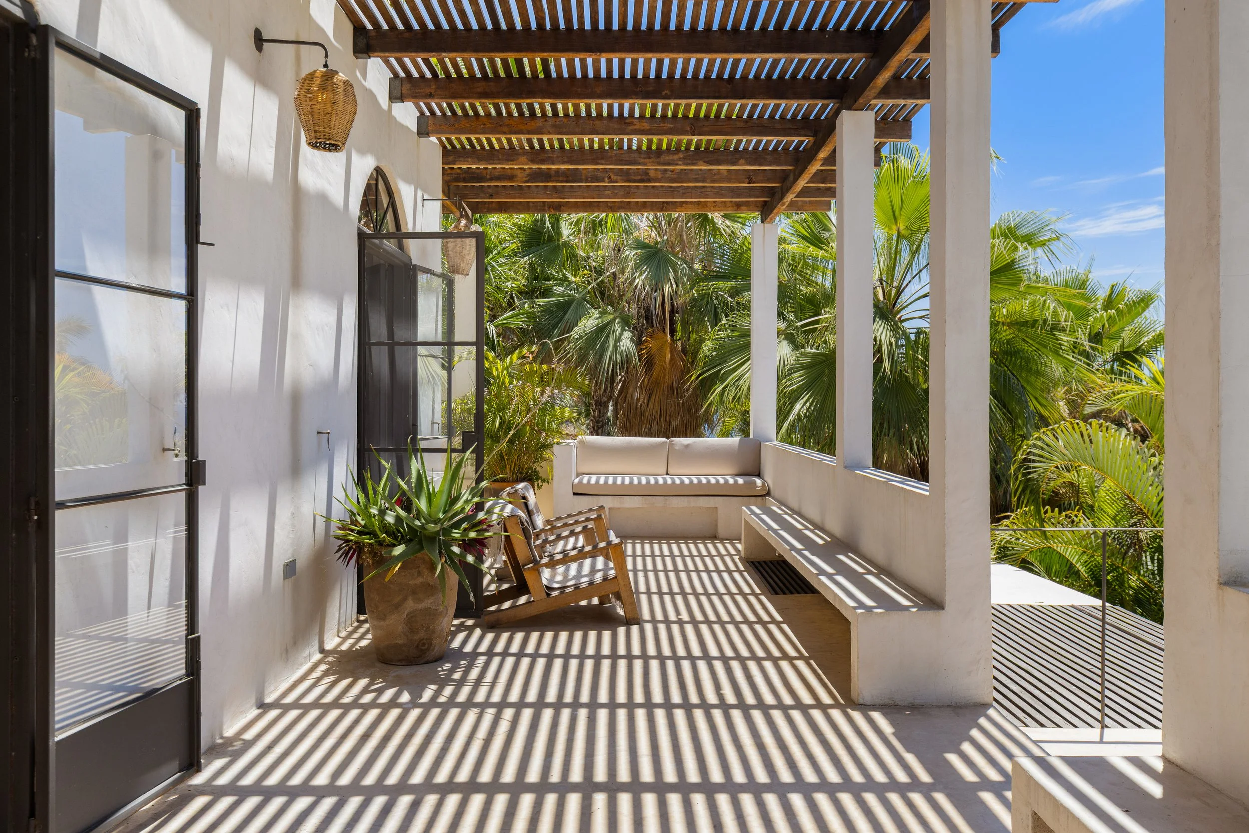 A balcony with white walls and a wooden pergola creating striped shadows on the floor and walls. There are chairs, a potted aloe vera plant, and lush green palm trees visible beyond the balcony.