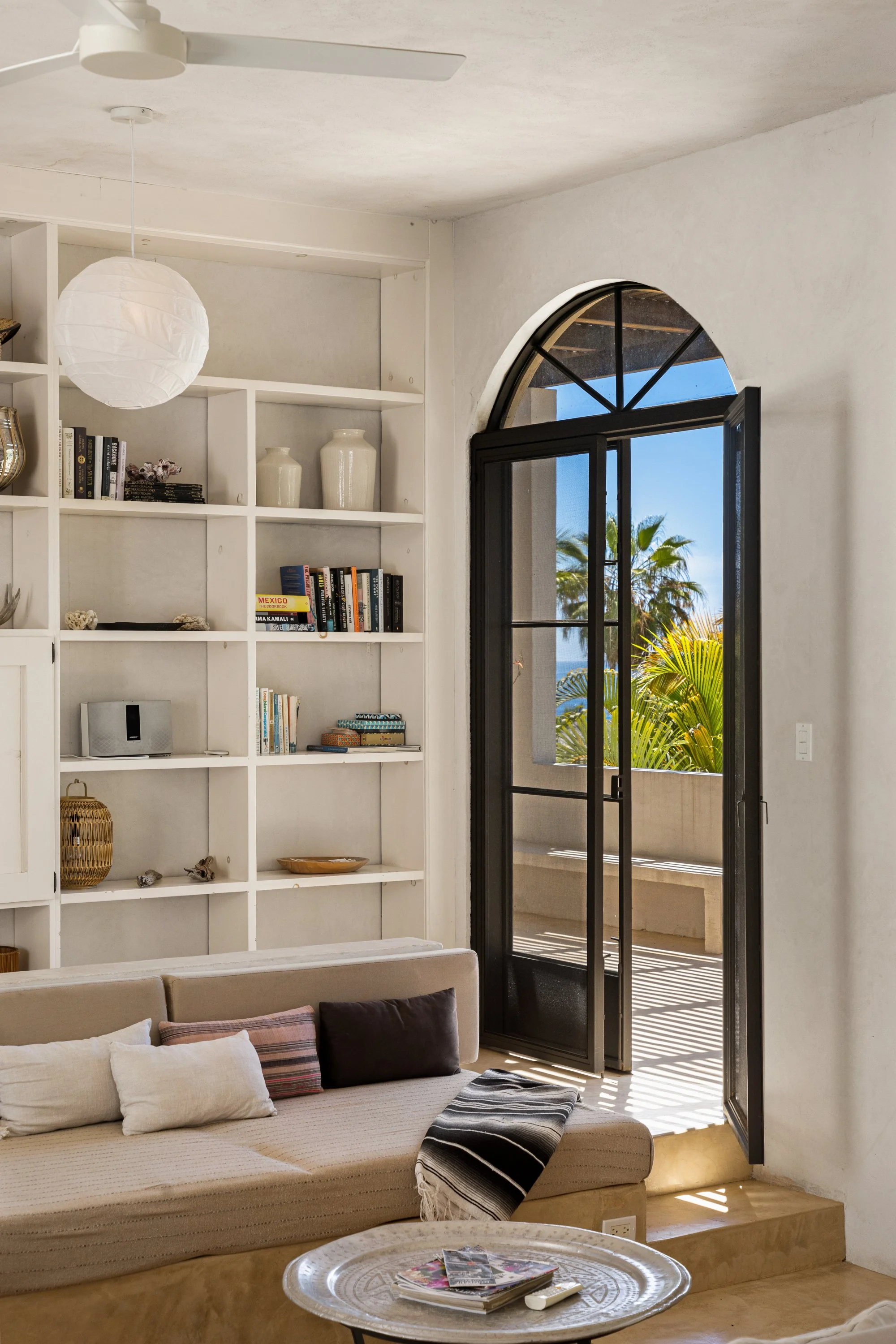 Living room with a beige sofa, throw pillows, and a round silver coffee table with magazines. There is a white wall with built-in shelves holding books and decorative items. An open black-framed glass door leads to a balcony with a view of palm trees