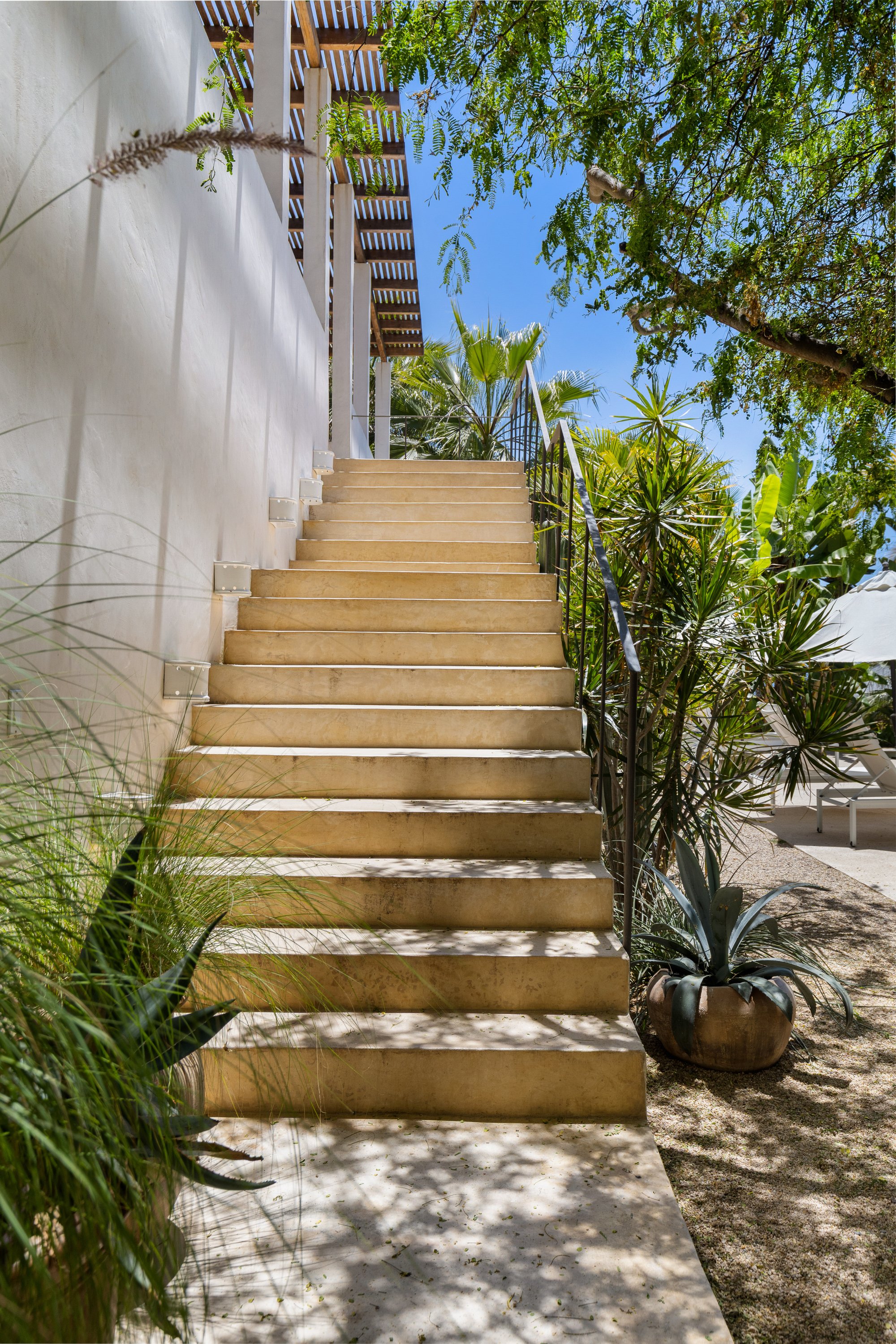Outdoor staircase made of beige stone with black metal handrails, surrounded by green plants and trees, leading up to a wooden platform and railing against a bright blue sky.