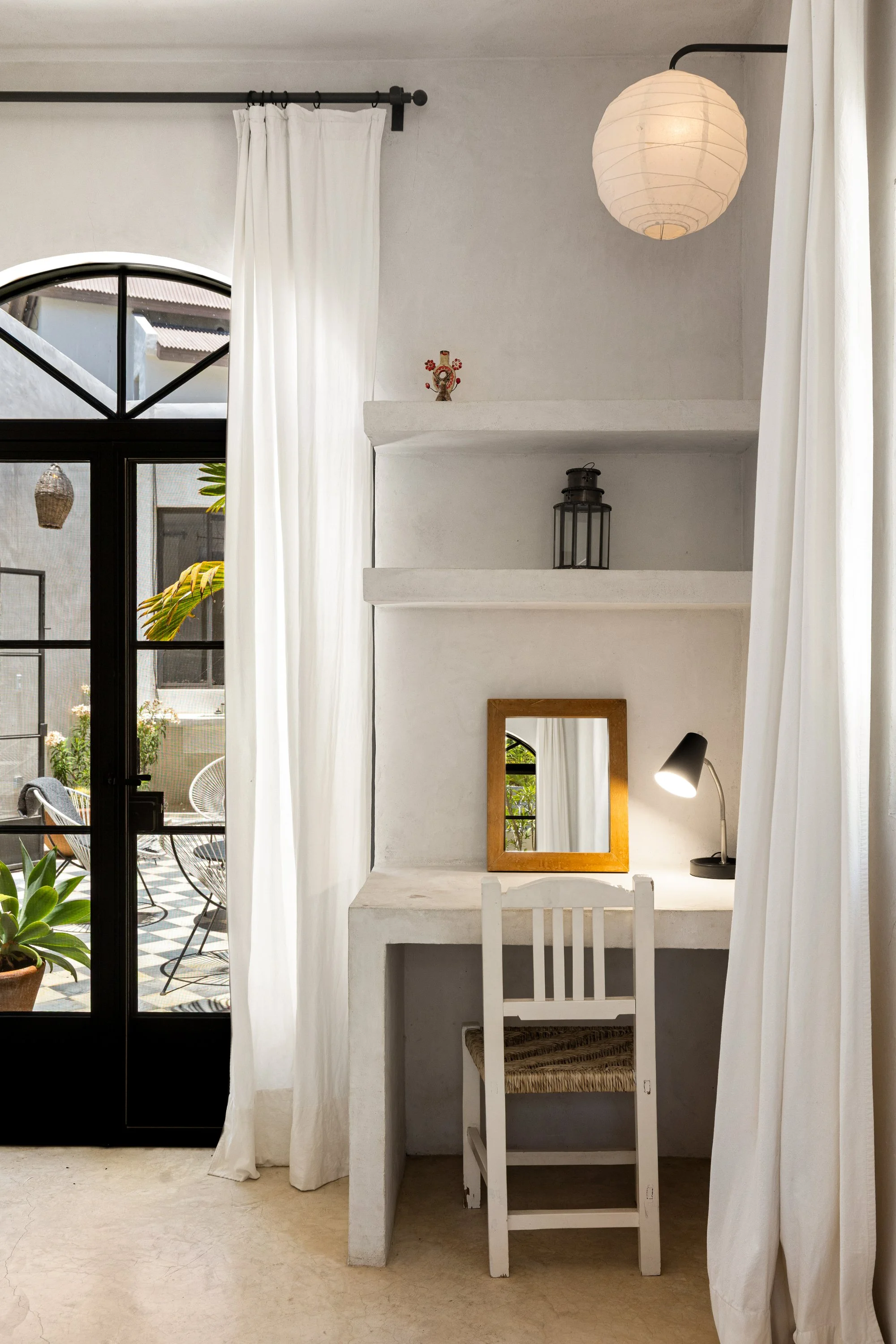 A cozy, minimalist interior featuring white curtains, a small white desk with a chair, a framed mirror, a black desk lamp, and decorative items on shelves. Through the glass door, an outdoor patio with chairs, a patterned floor, and greenery is visib