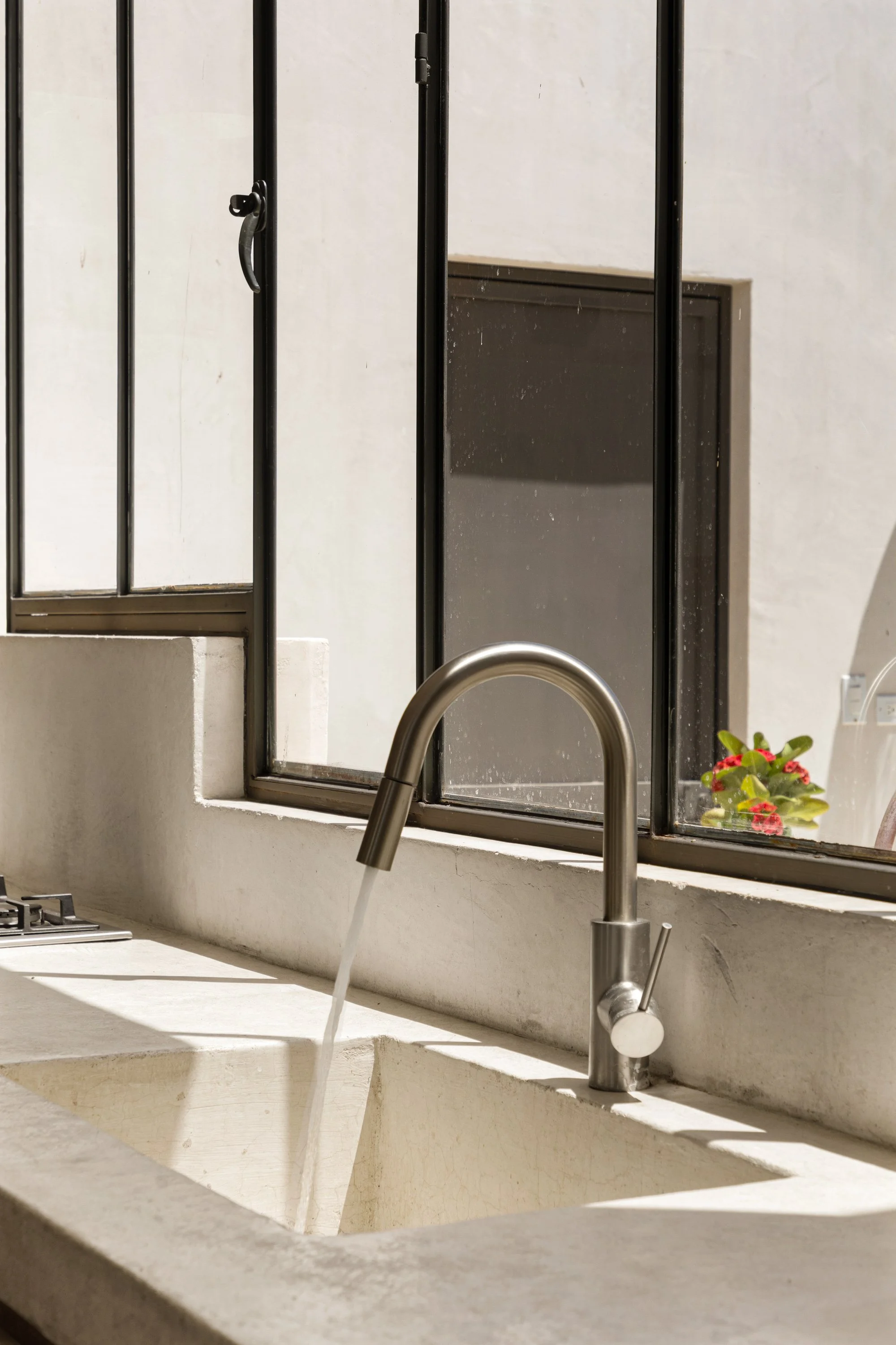 A kitchen sink with a silver faucet, running water, a window with a black frame, a small potted plant with red flowers, and a vent in the wall.