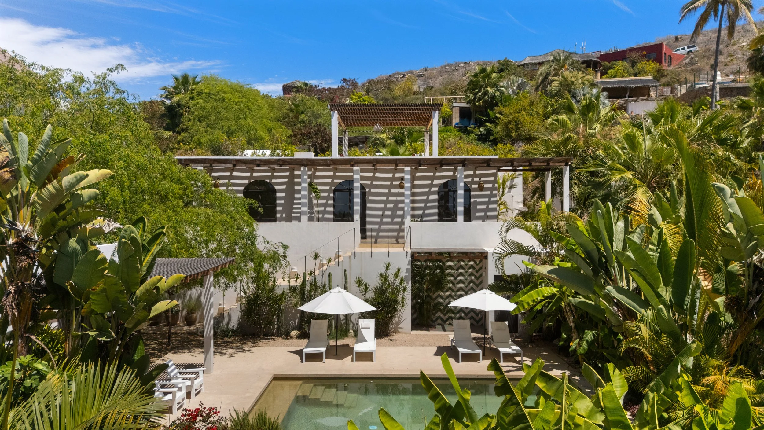 A picturesque scene of a modern white house surrounded by lush green tropical plants and trees, with a small pool and lounge chairs in the foreground, set against a hillside with additional houses and a bright blue sky with scattered clouds.