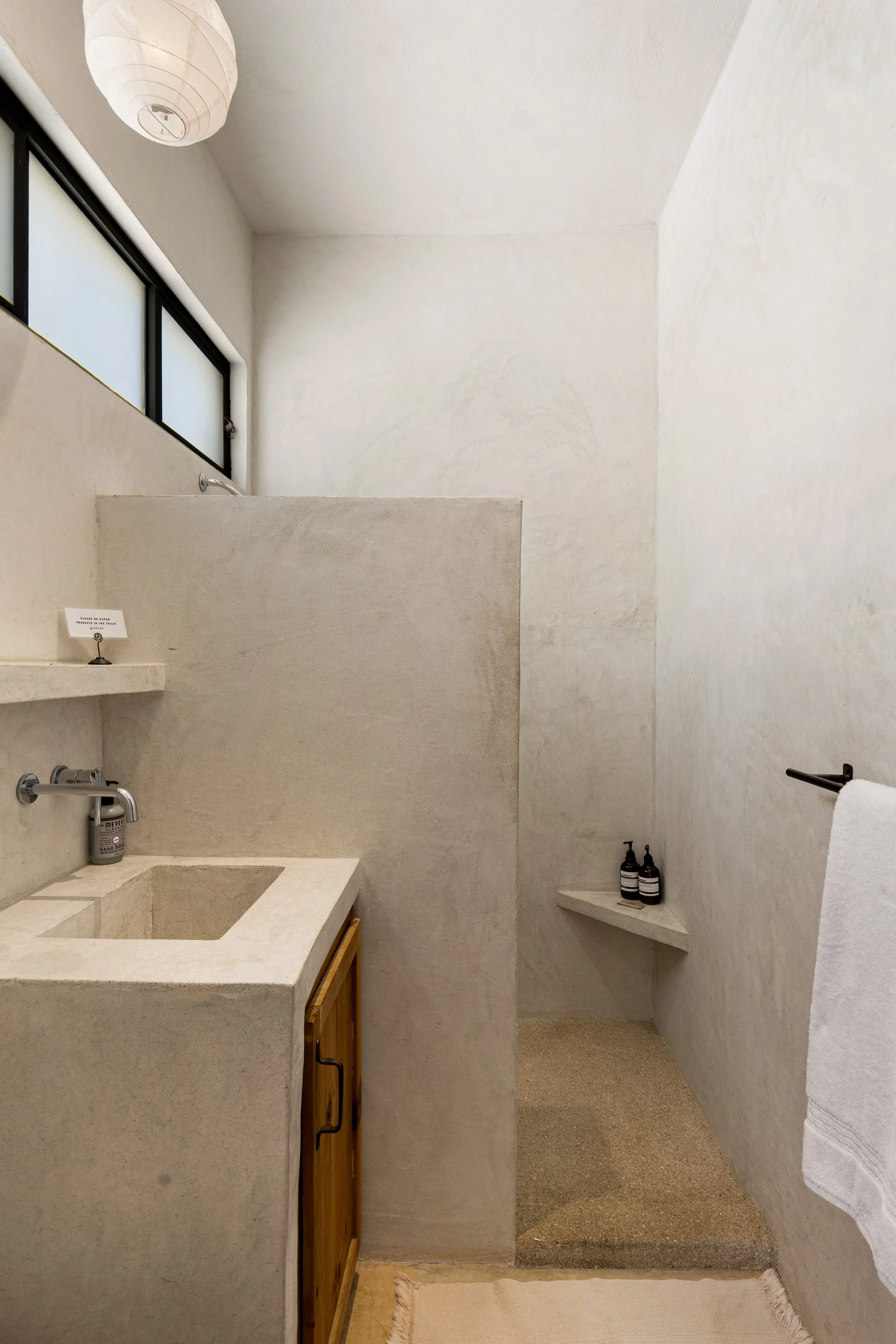 A minimalist bathroom with white textured walls, a concrete sink with a wooden cabinet below, a small corner shelf with two black bottles, a window with frosted glass, and a white paper lantern ceiling light.