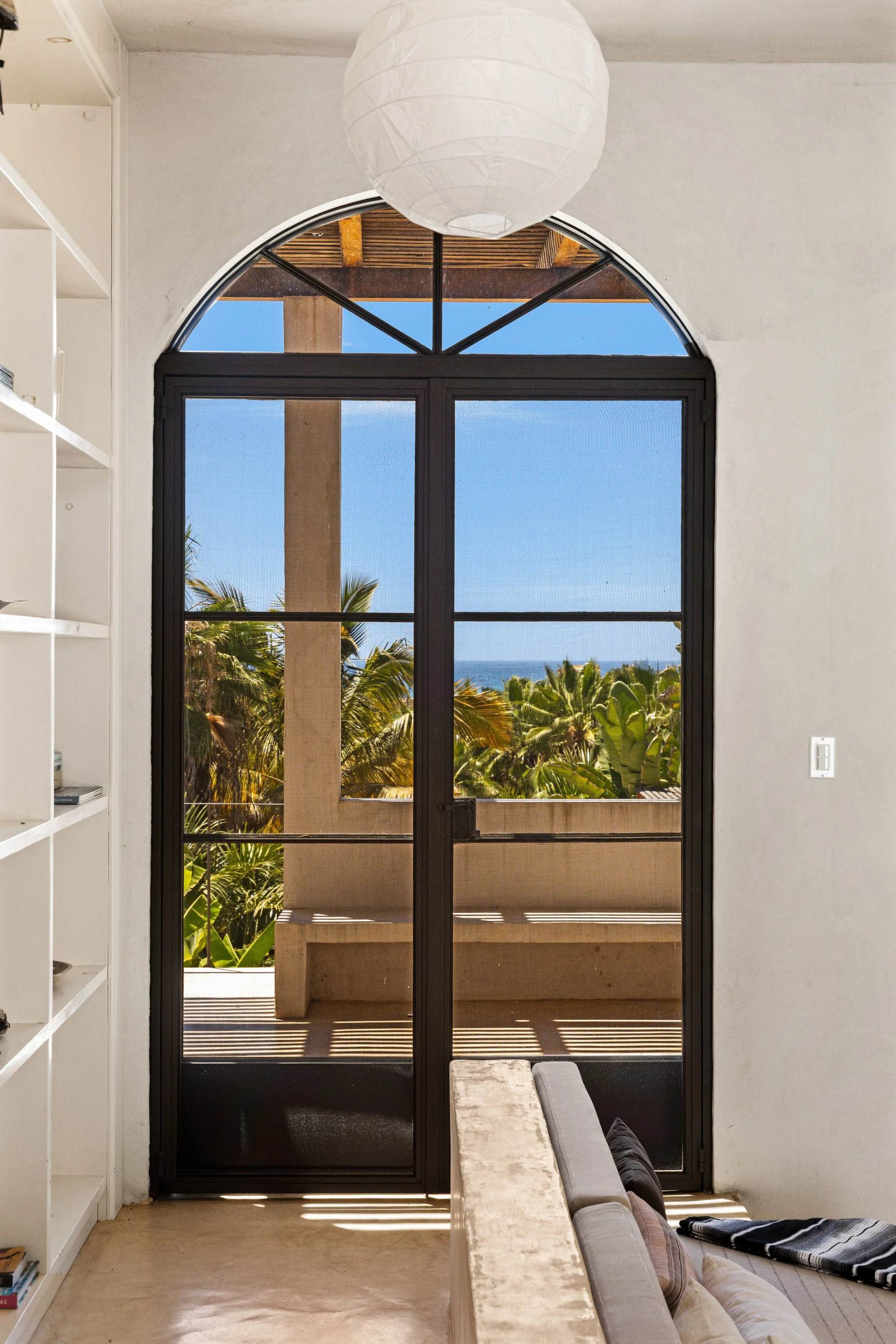 View through a black-framed glass door showing a balcony with a sitting bench, tropical plants, and the ocean in the distance under a partly cloudy sky.