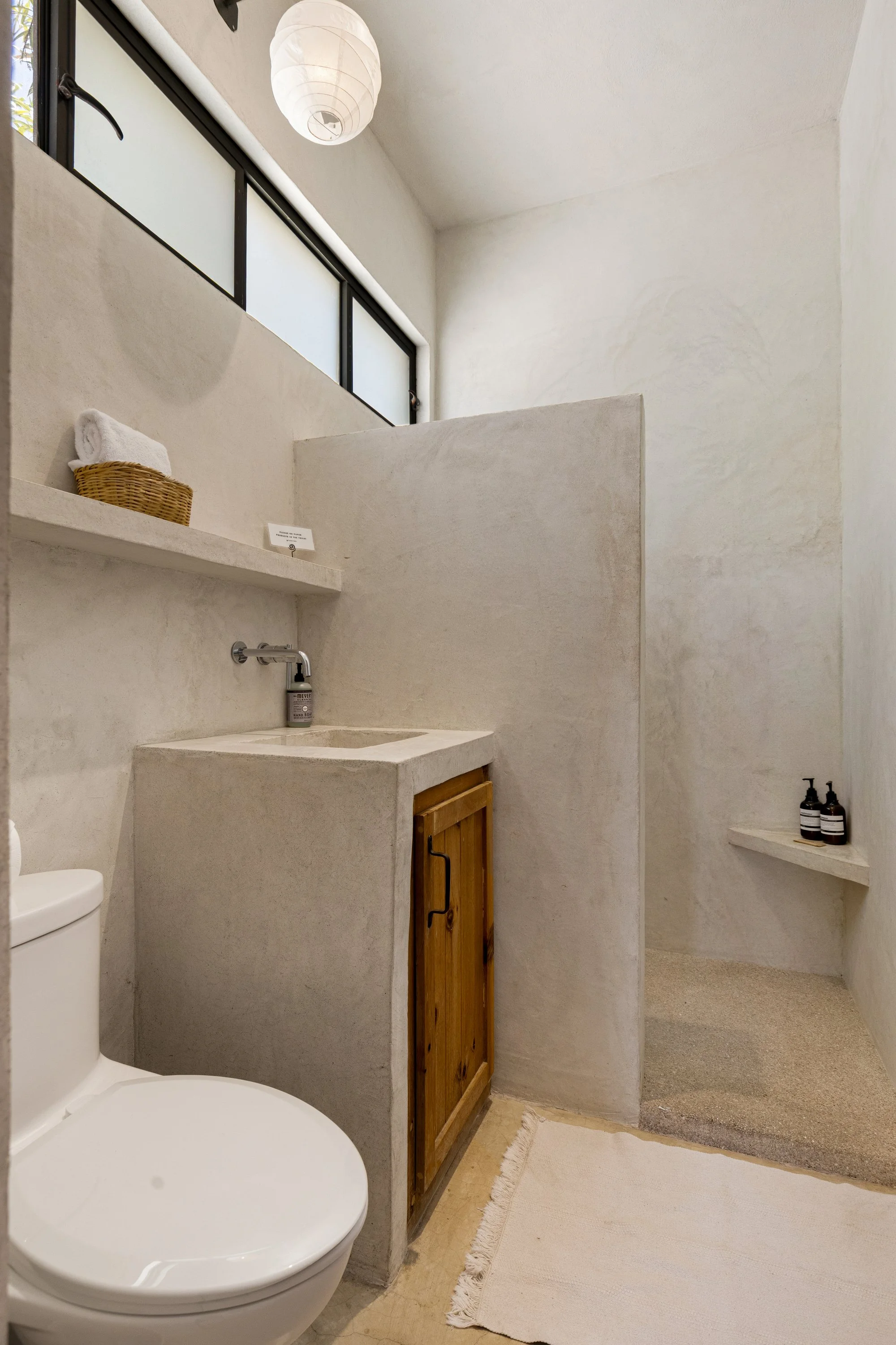 Minimalist bathroom with white textured walls, a small sink with a rustic wooden cabinet, a white toilet, a floating shelf with black soap dispensers, a basket of rolled towels, large frosted window, and paper lantern light, with a soft beige rug.