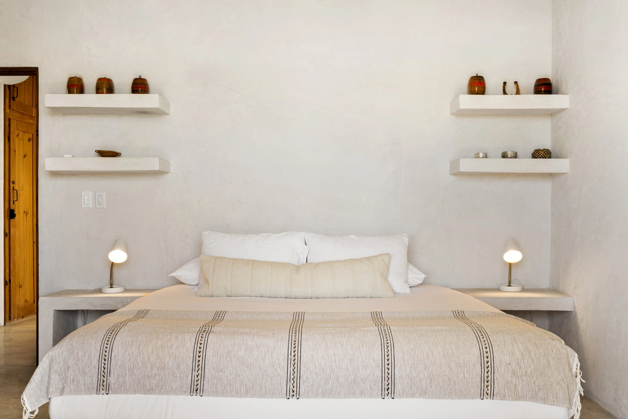A minimalist bedroom with a white bed, beige throws, two white pillows, and two small bedside lamps with curved stands. The wall behind the bed has two white floating shelves displaying decorative vases, figurines, and bowls. A wooden door is partial