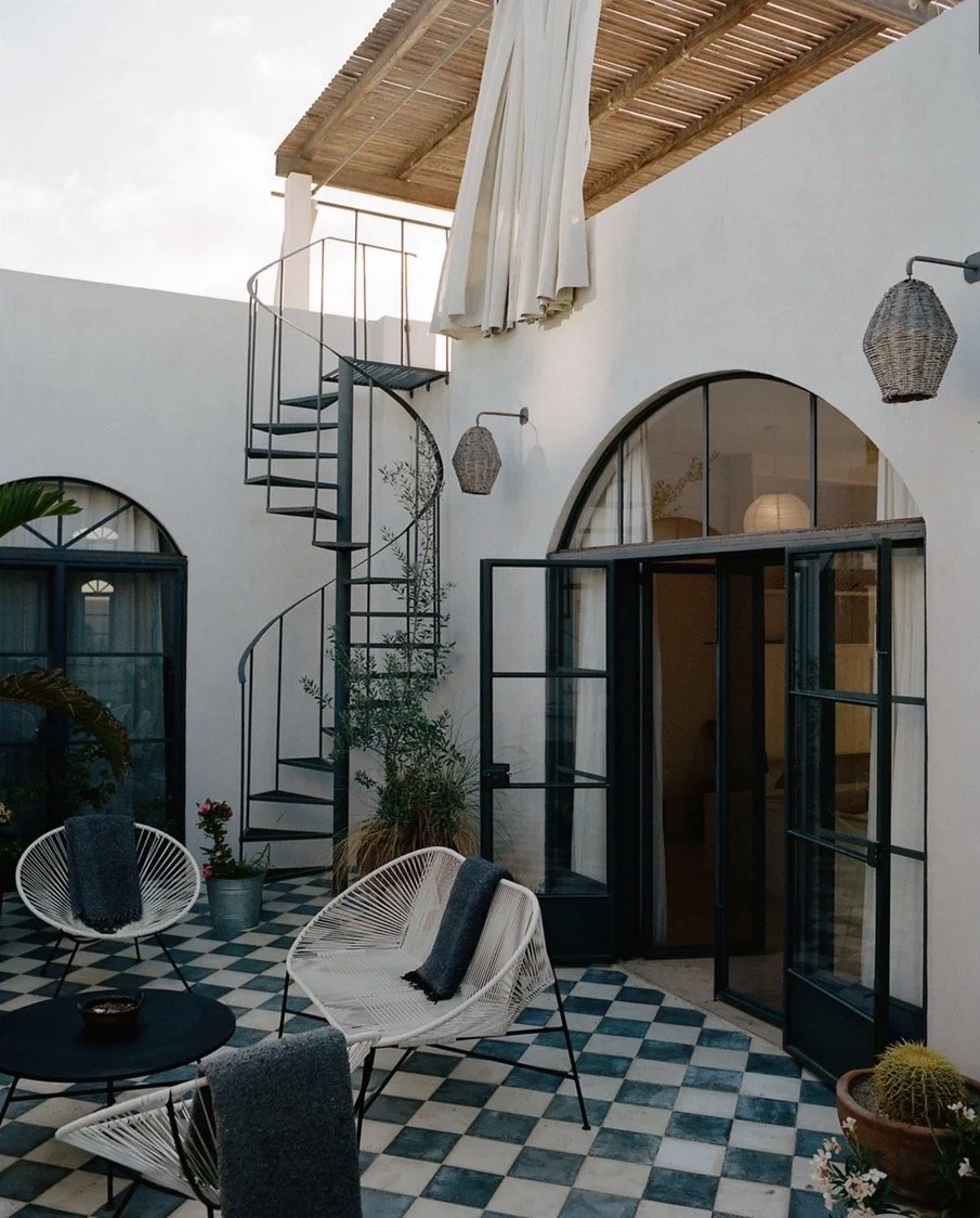 Outdoor patio with checkered tile floor, white and black chairs with dark towels, plants, black metal spiral staircase, large arched glass doors, white walls, wooden roof, and wicker wall lamps.