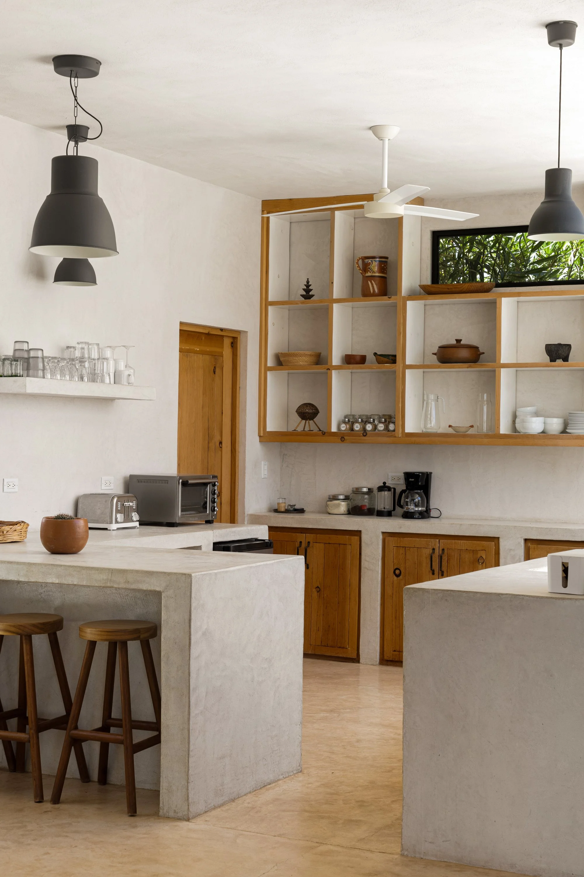 A modern kitchen with beige countertops, wooden cabinets, open shelving with decorative items, white walls, and two black pendant lights hanging from the ceiling. There is a window with greenery outside and a white ceiling fan.