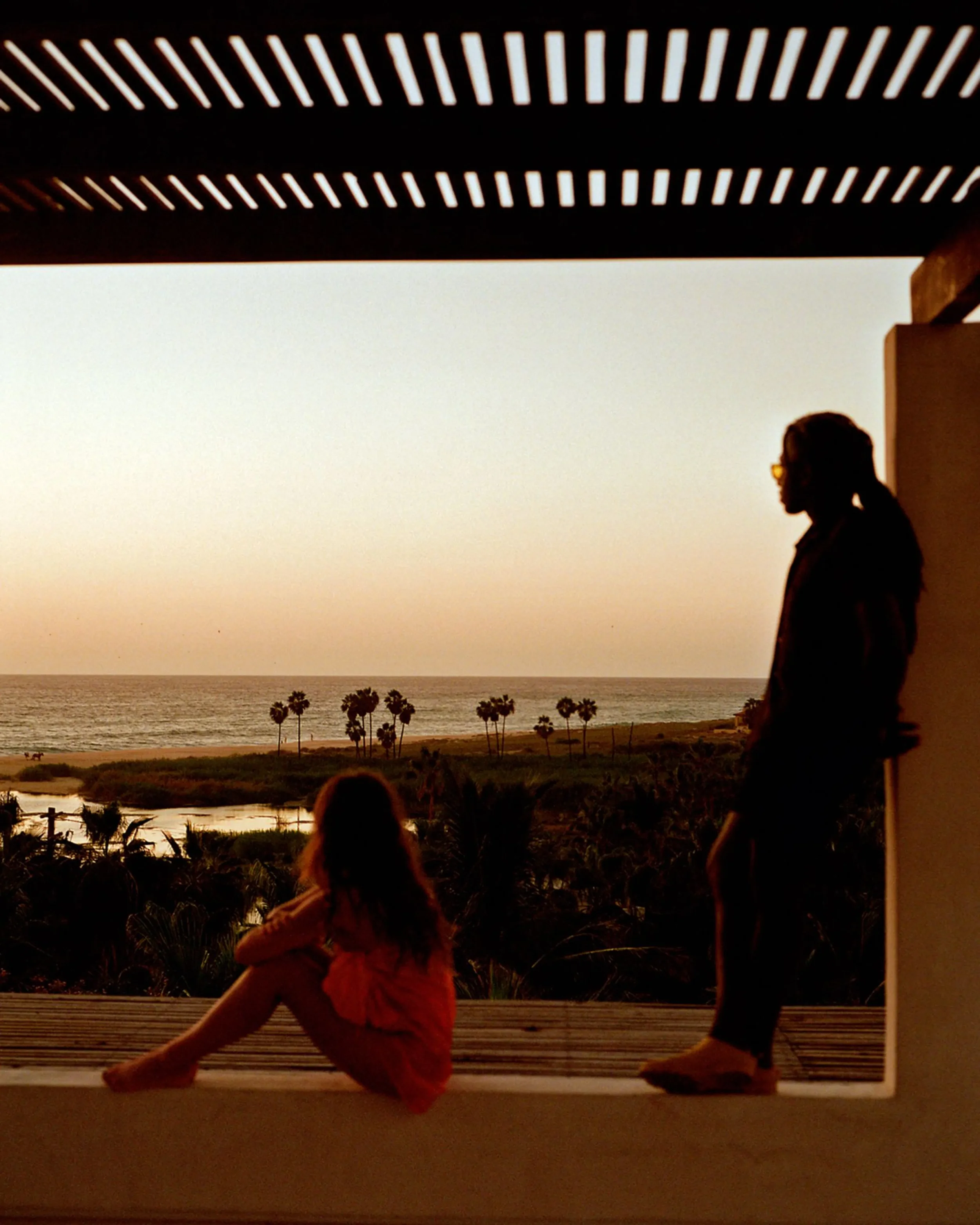 Children playing on a window ledge overlooking a sunset over the ocean with palm trees in the background.