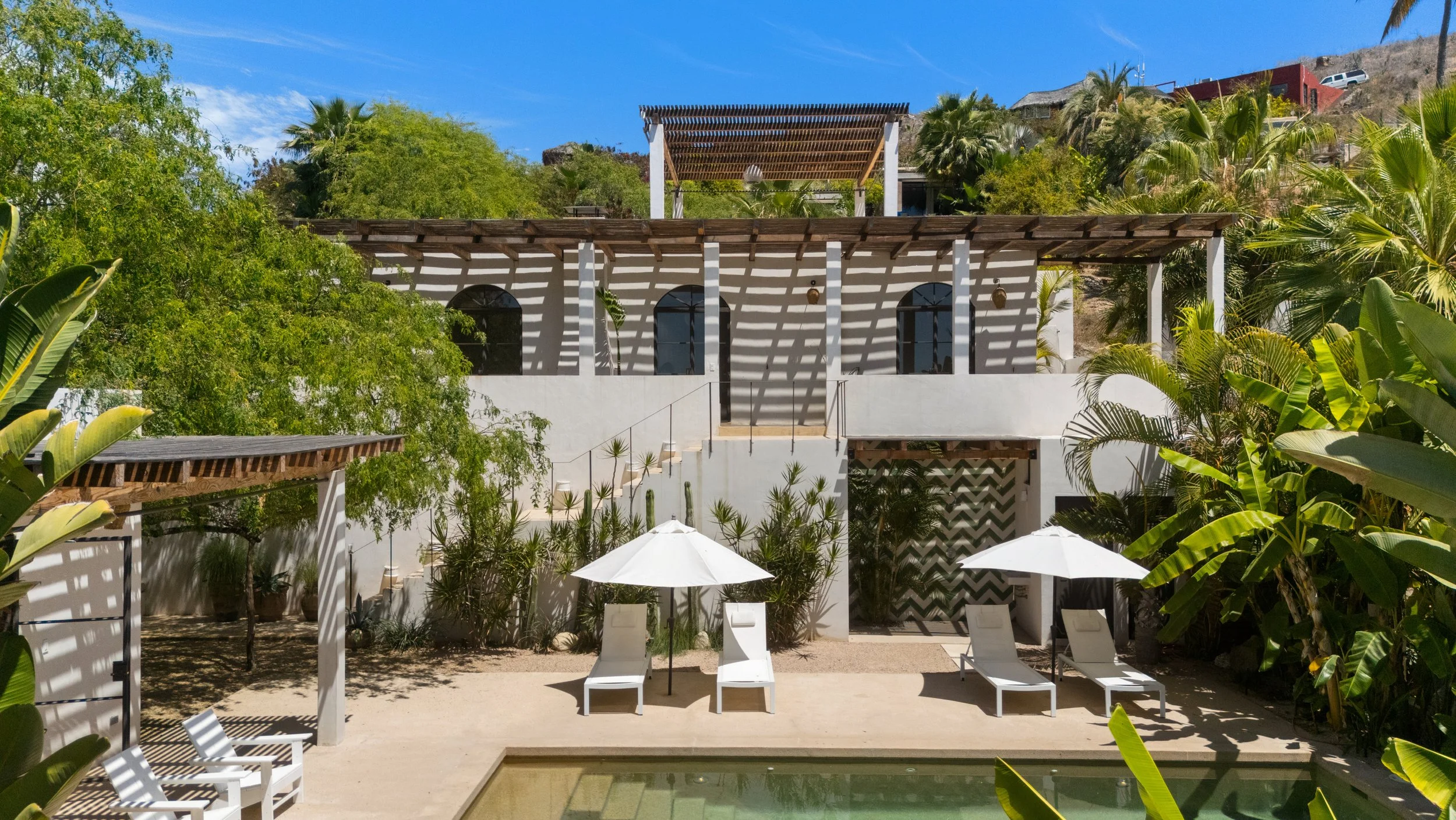A modern white building with arched windows surrounded by lush green trees and palm trees, with outdoor lounge chairs and umbrellas in the foreground, under a clear blue sky.