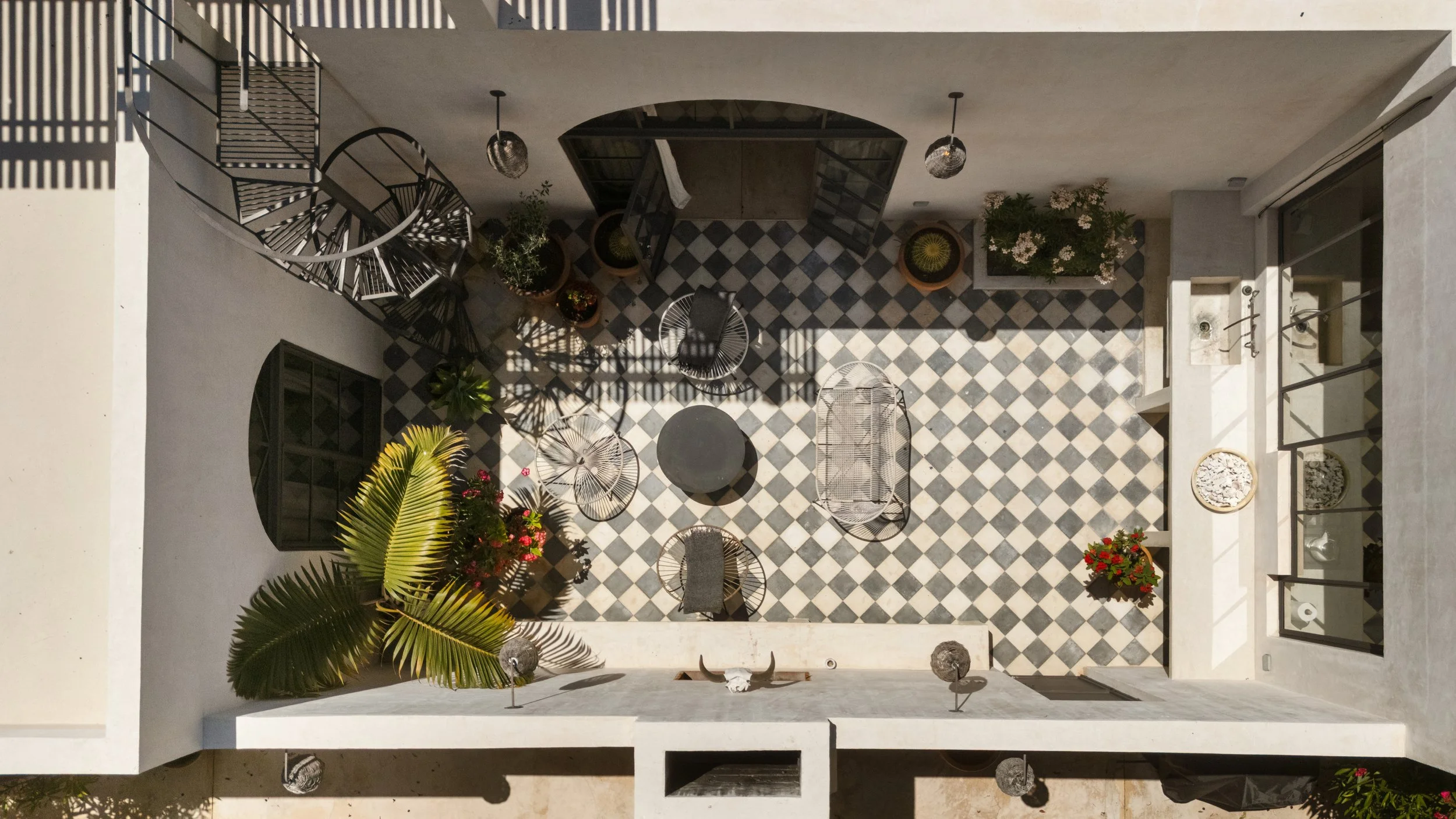 A rooftop patio with checkered black and white tile flooring, outdoor furniture including chairs and a round table, potted plants, and decorative elements like hanging lanterns and sculptures, with sunlight casting shadows.