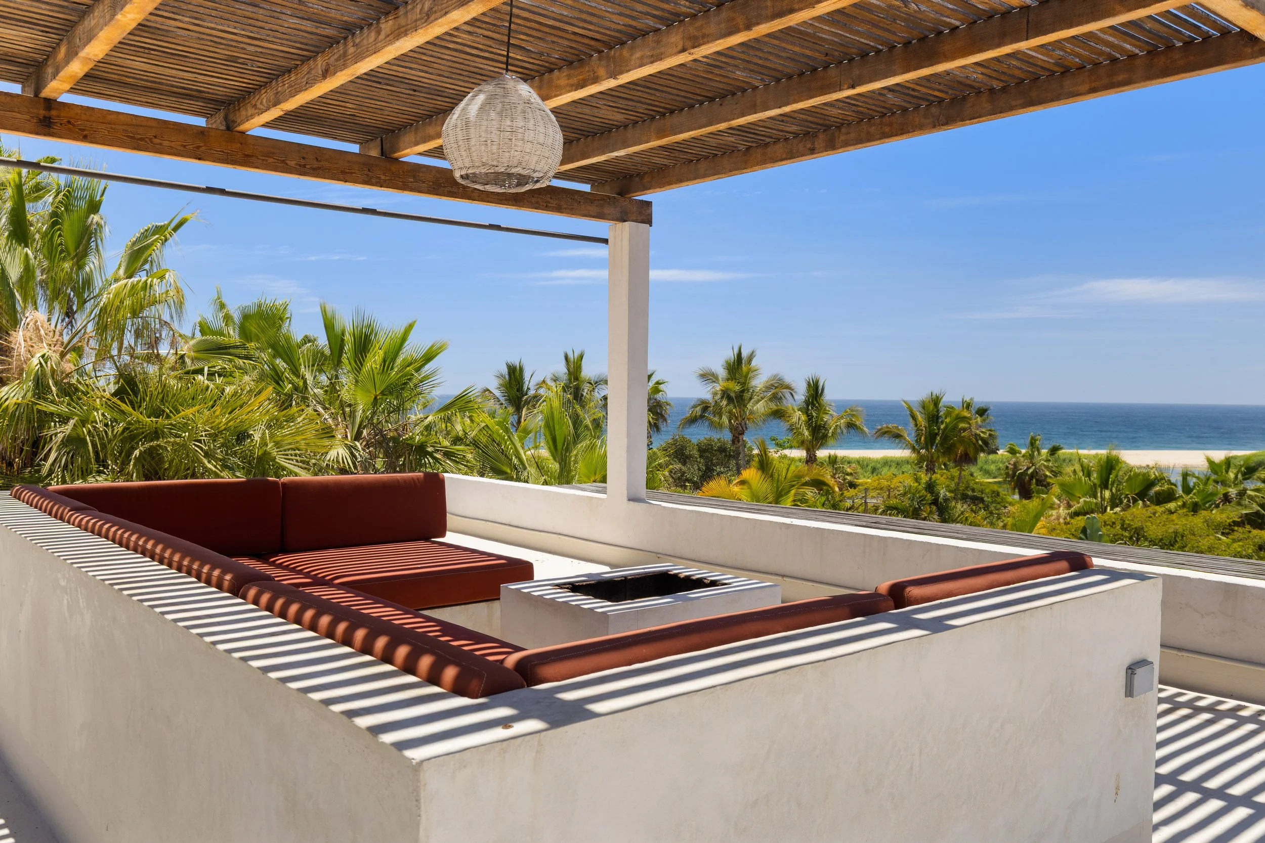 Outdoor terrace with built-in seating and a shaded pergola, overlooking a tropical landscape with palm trees and the ocean in the distance.