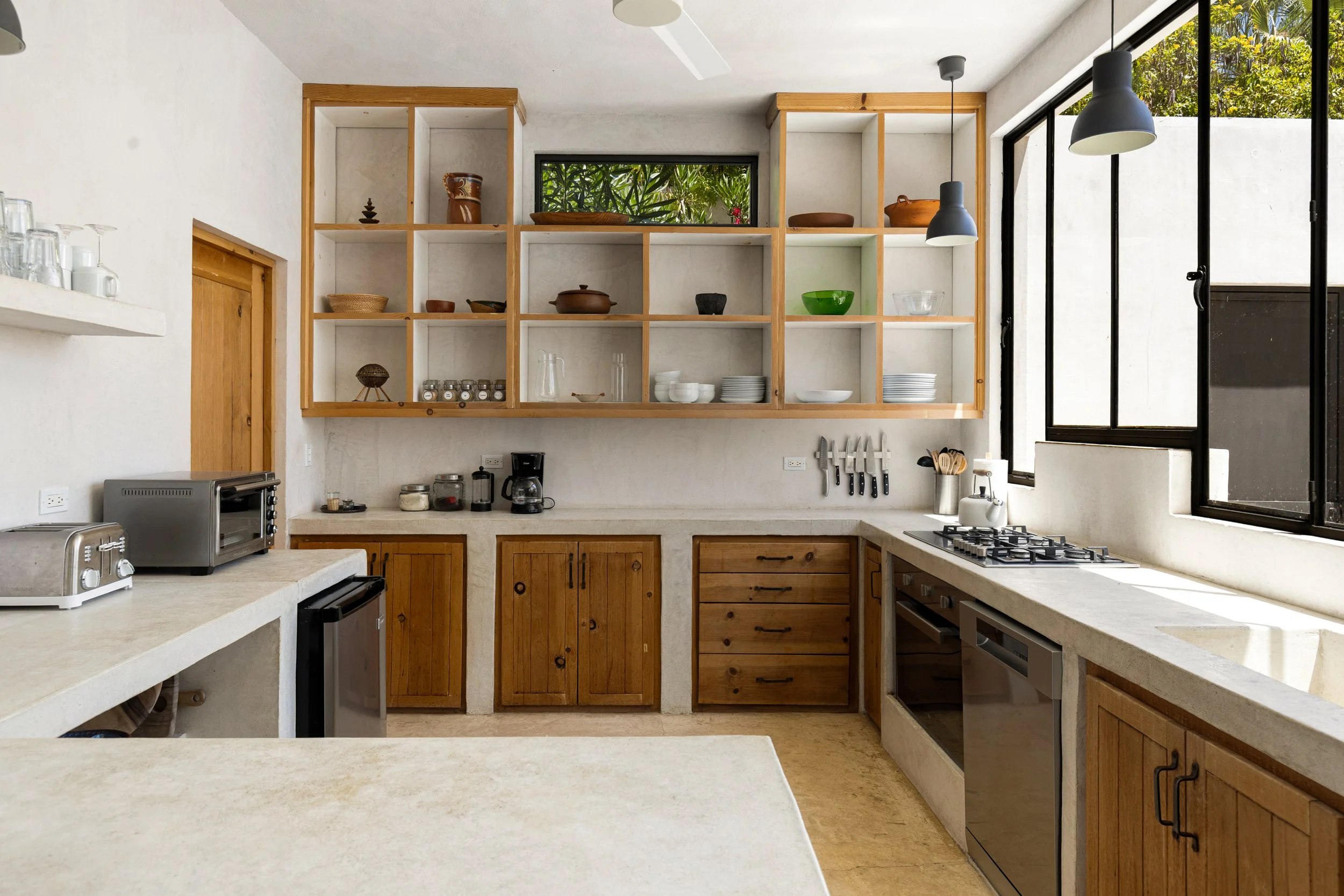 Bright kitchen with open wooden shelving containing dishes, bowls, and pots, white countertops, black framed large windows, and pendant lights, with a view of greenery outside.