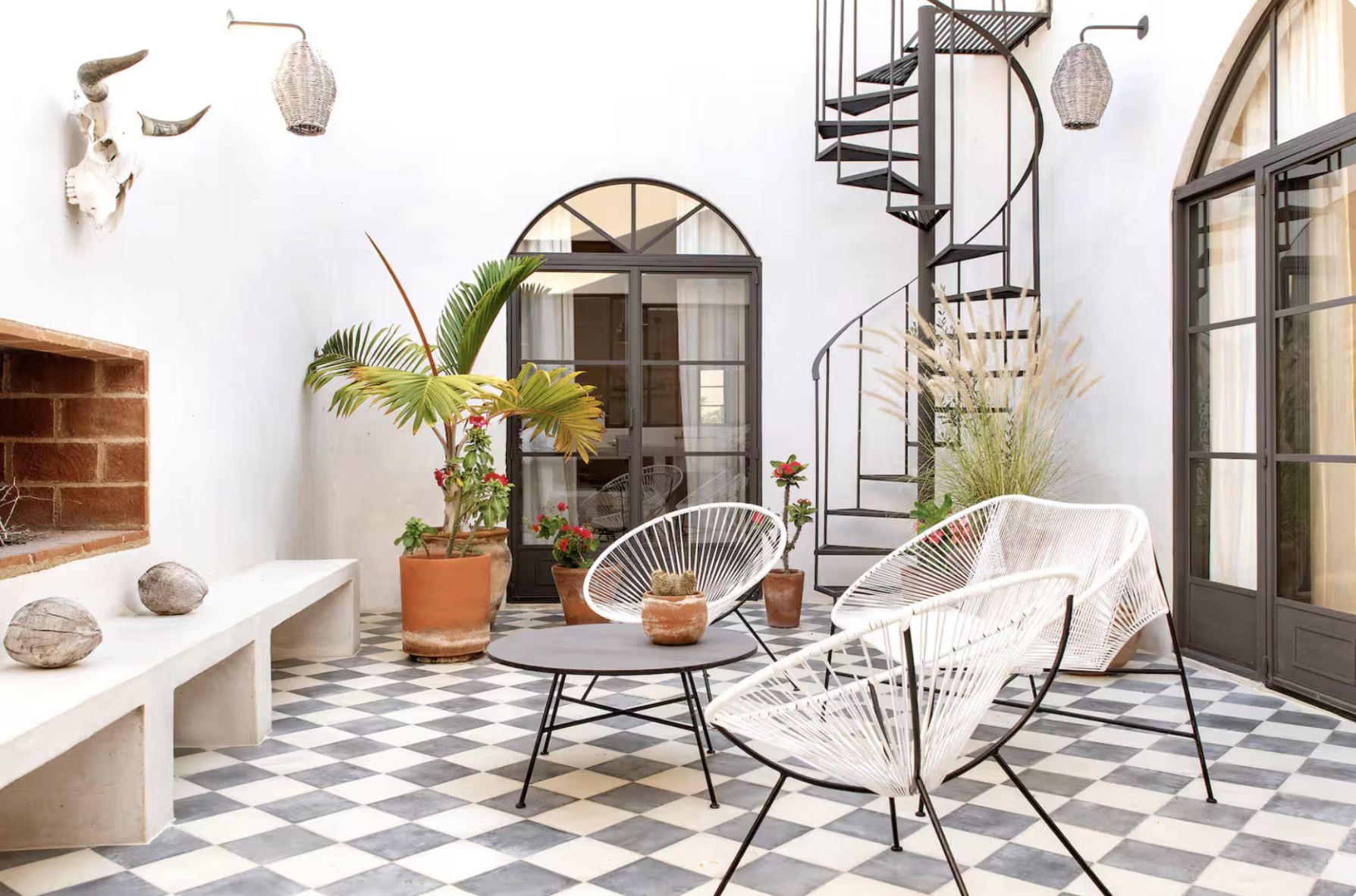 Indoor patio with white walls, black and white checkered tile floor, and modern furniture including white wire chairs around a small round table. Decor includes potted plants, a mounted animal skull with horns, and woven pendant lights. There is a spiral staircase and large windows with curtains.