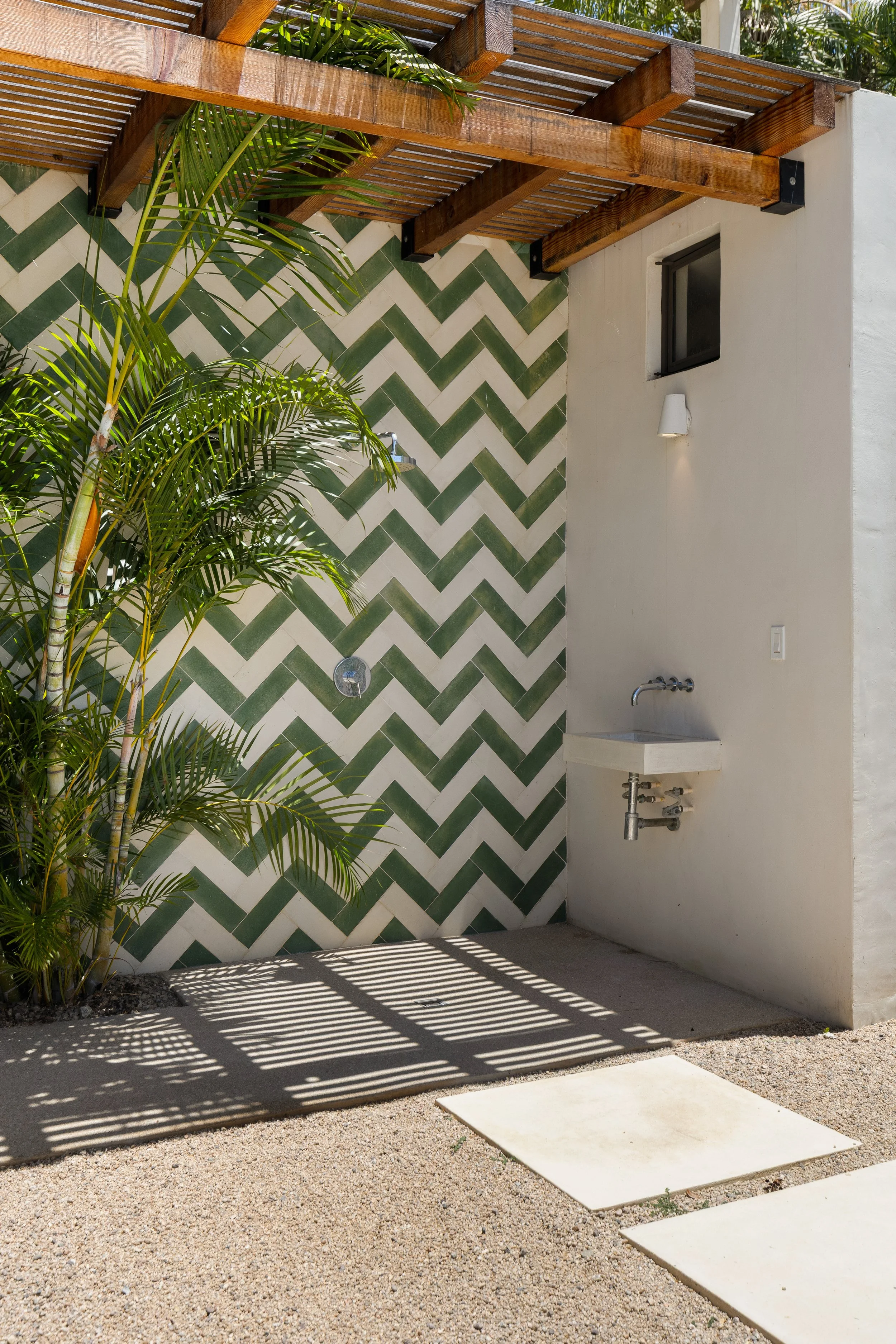 Outdoor space with a white wall featuring a black-framed window, a small wall-mounted white light, and a concrete sink below the window. The wall is decorated with green and white chevron tiles. There is a wooden pergola above, casting shadows on the gravel ground, and a potted palm plant in the left foreground.