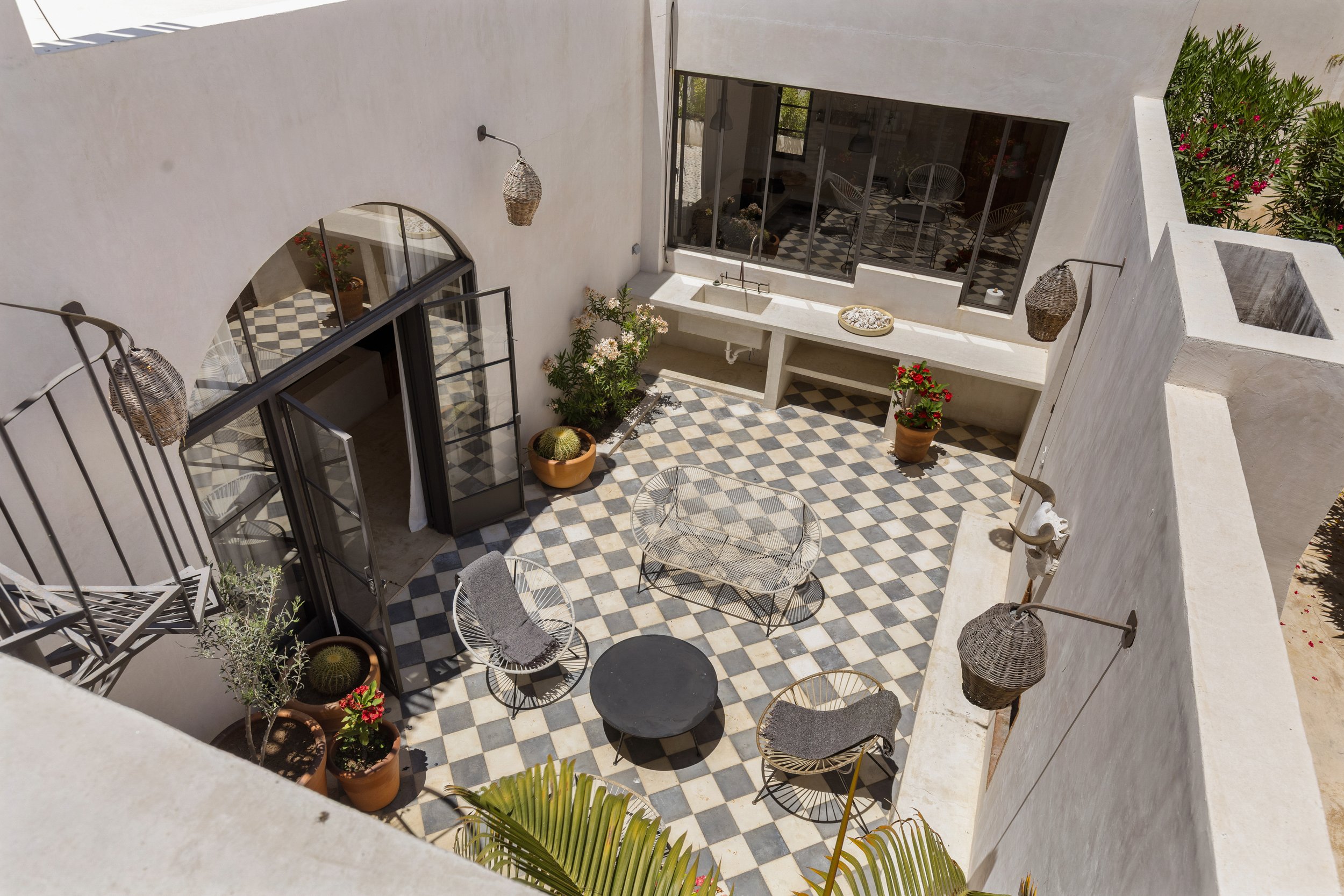An outdoor patio with black and white checkered tile flooring, patio furniture including a round table with chairs and a bench, potted plants, and large windows reflecting interior space. The patio is enclosed by white stucco walls.