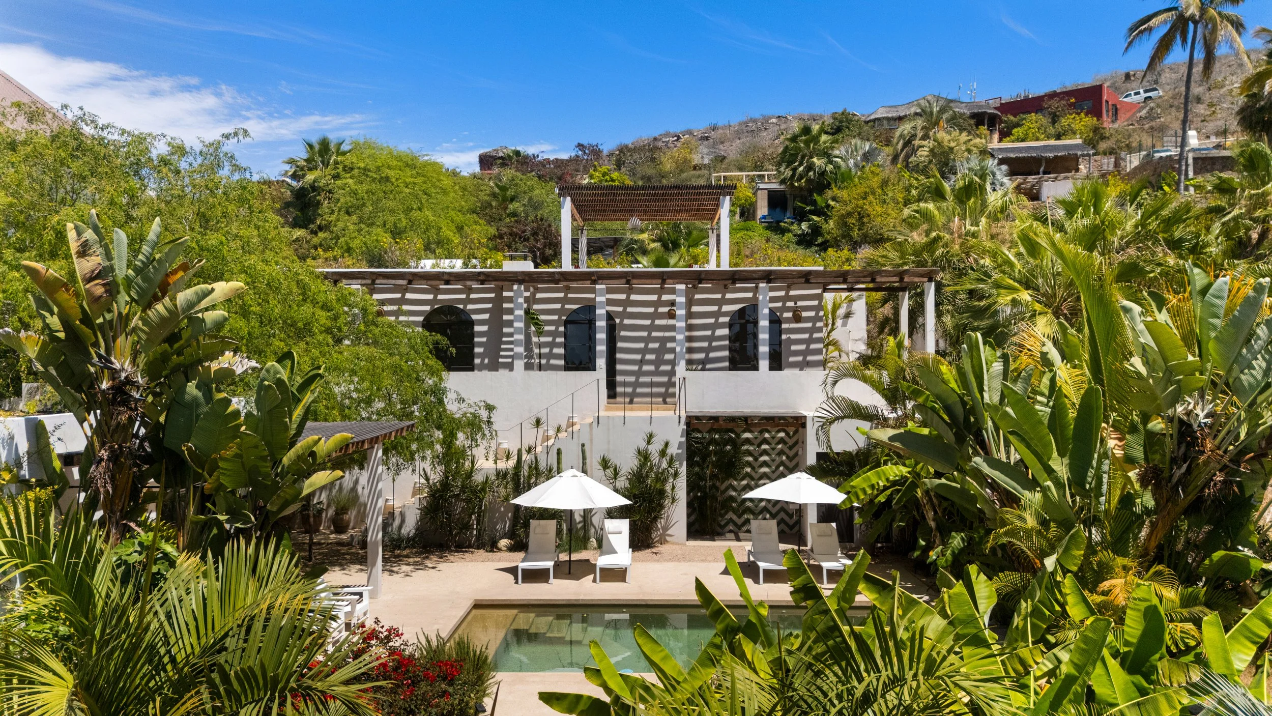 A luxurious white villa with a pool, surrounded by lush green tropical plants and palm trees, with a hillside in the background under a bright blue sky.