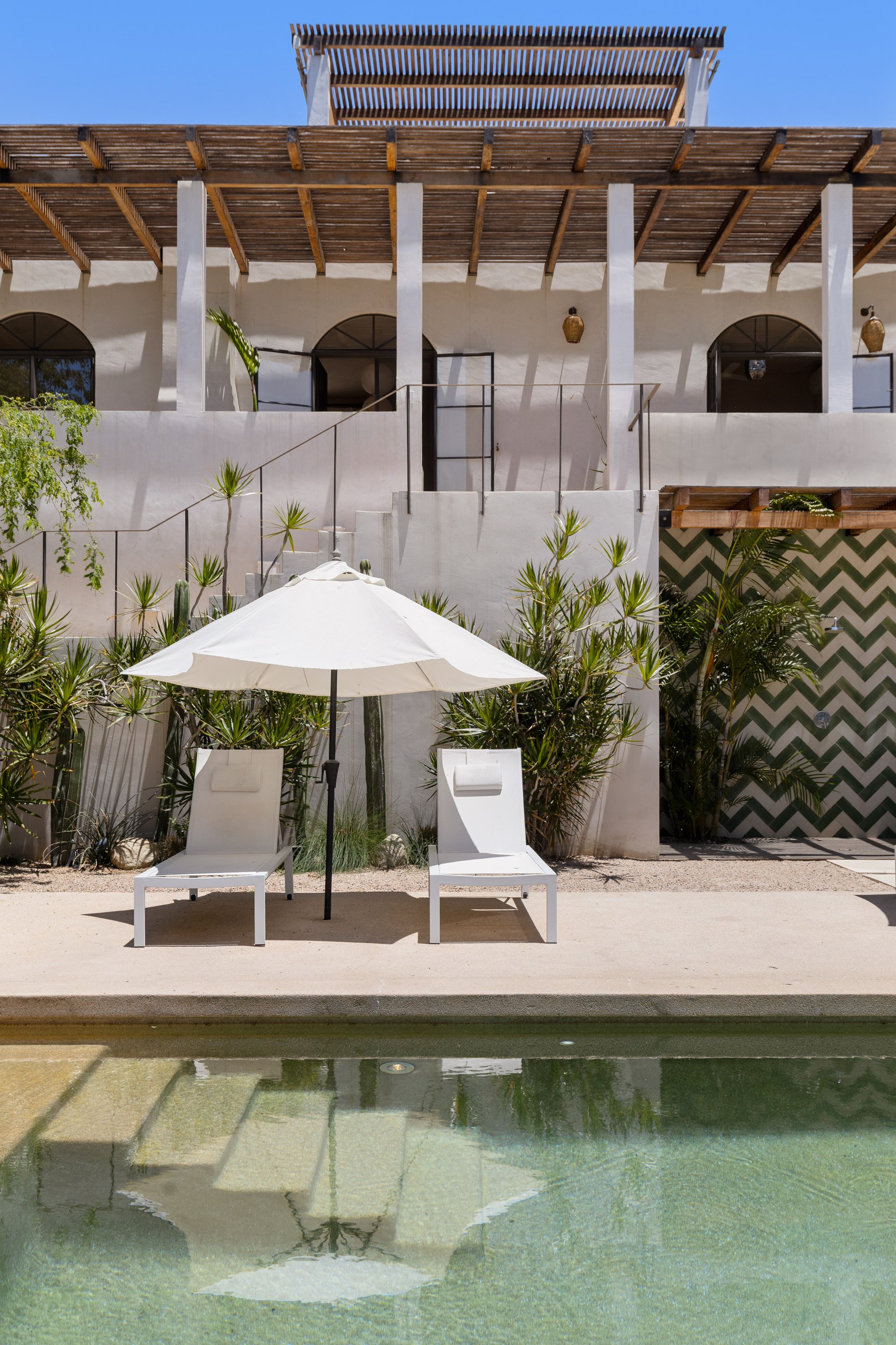 The image shows a modern white building with two levels, wooden pergolas, and arched windows. There are two white lounge chairs with an umbrella in front of the building and a small pool or water feature in the foreground, reflecting parts of the bui