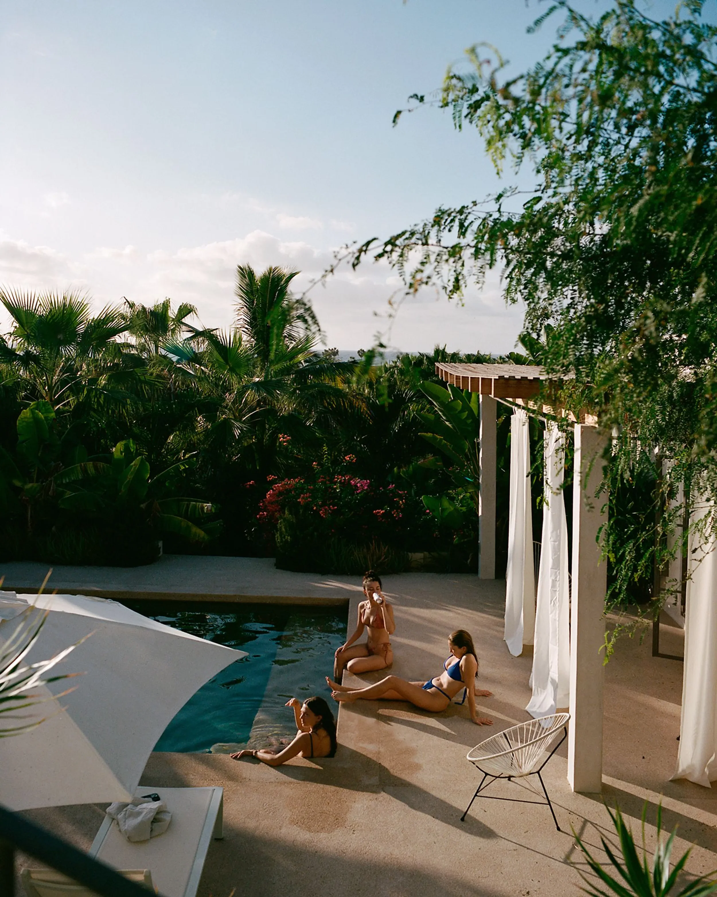 Four women relaxing by a swimming pool on a sunny day. One woman is sitting on the pool's edge, drinking from a cup, while another is lying on a towel sunbathing. Two women are sitting at the poolside, one leaning back and the other sitting with her legs stretched out. The scene is surrounded by lush greenery and a wooden pergola with white curtains.