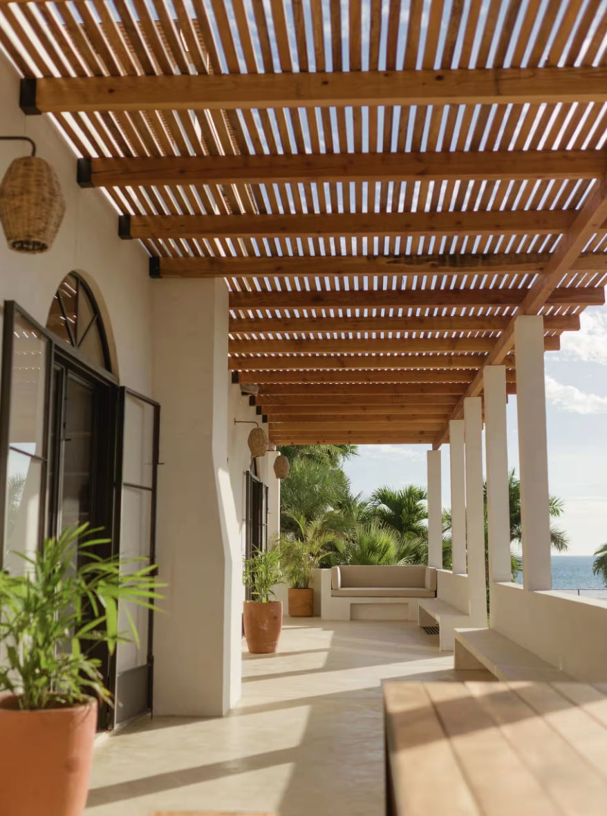 Bright outdoor covered patio with white stucco walls, wooden slat roof, and tropical plants. There is a built-in bench with beige cushions and potted plants, overlooking palm trees and ocean views in the distance.