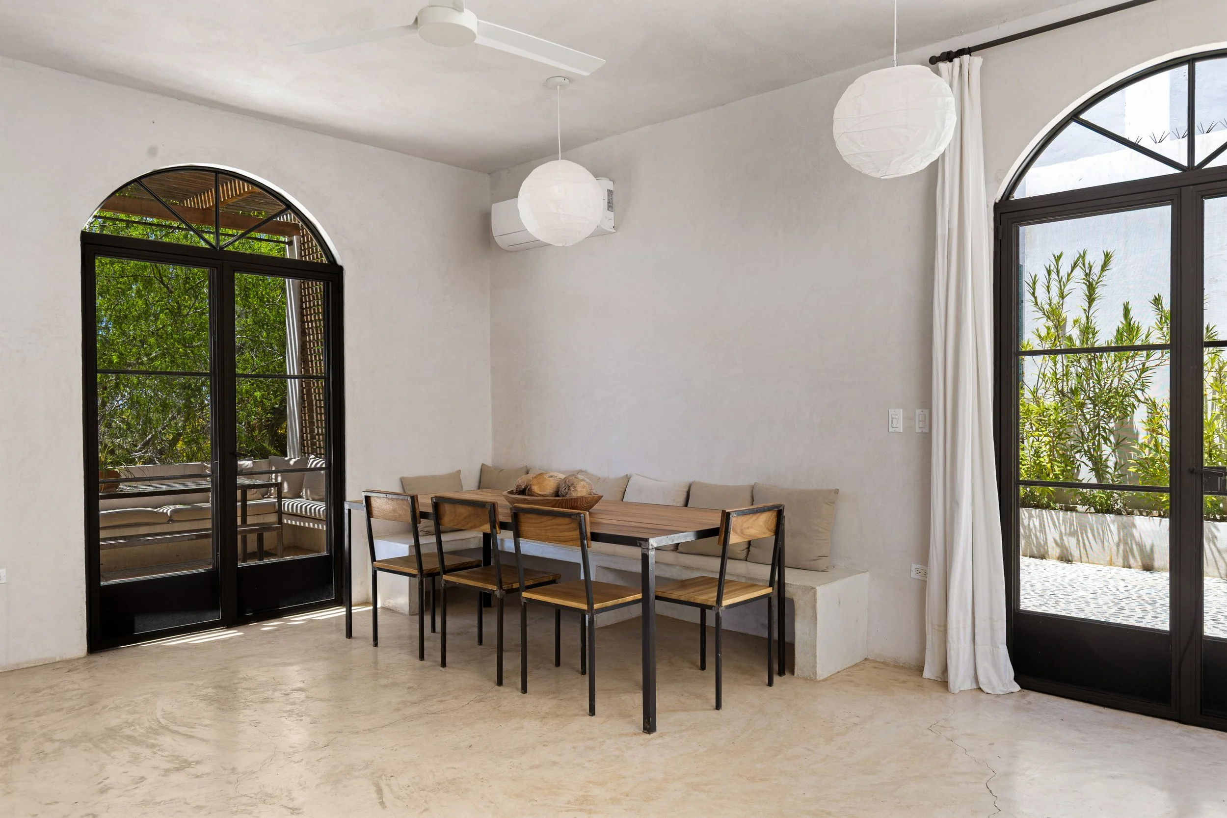 A bright dining area with large arched windows, white curtains, and a white ceiling fan. There is a wooden dining table with black metal chairs, and a beige sofa along the wall. Two white paper lantern pendant lights hang from the ceiling, and greene