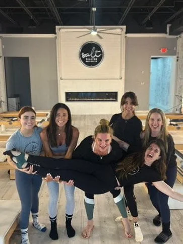 A group of five women posing and holding another woman horizontally in a dance studio.