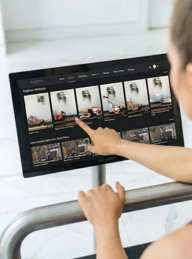 Person using a touchscreen monitor to select Pilates workout videos with images of people exercising on a Pilates reformer machine.