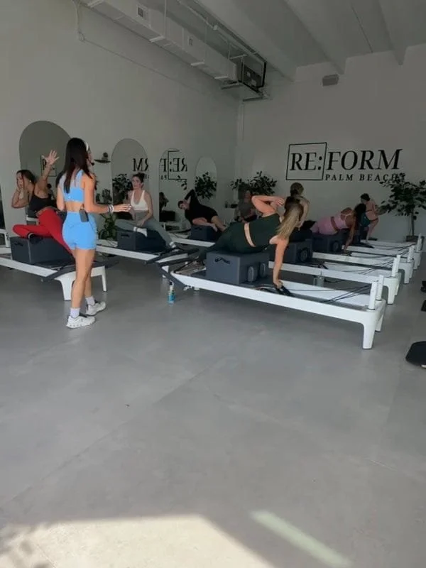 People participating in a Pilates class on reformer machines in a fitness studio with white walls and minimal decor.