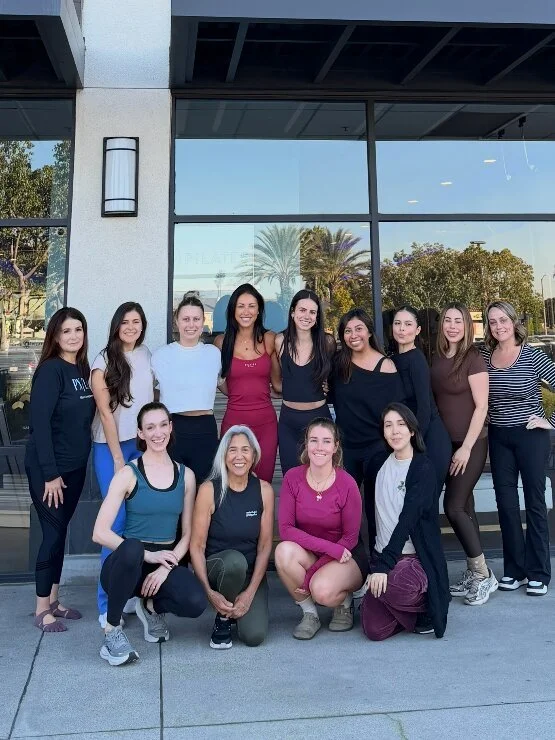 Group of women posing outdoors in front of a building with large windows and palm trees reflection.