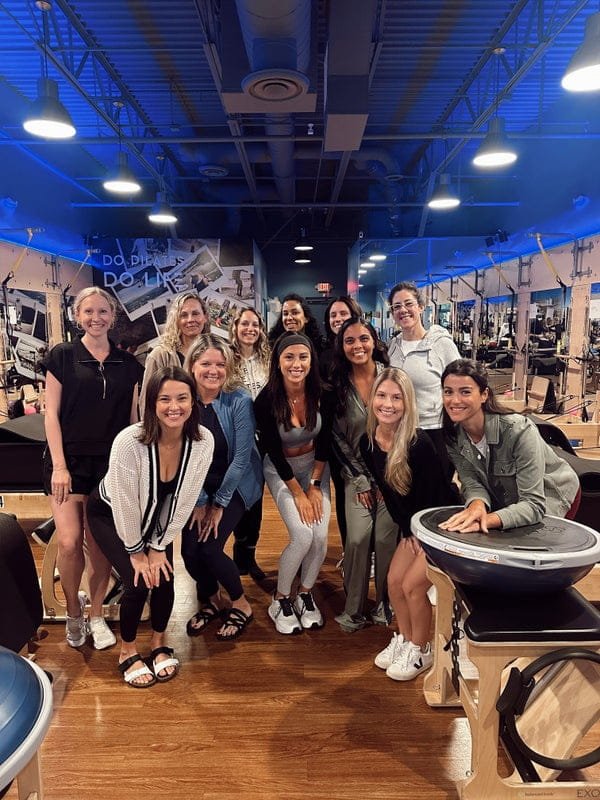 Group of thirteen women posing for photo in fitness studio with mirrors, exercise equipment, and blue lighting.
