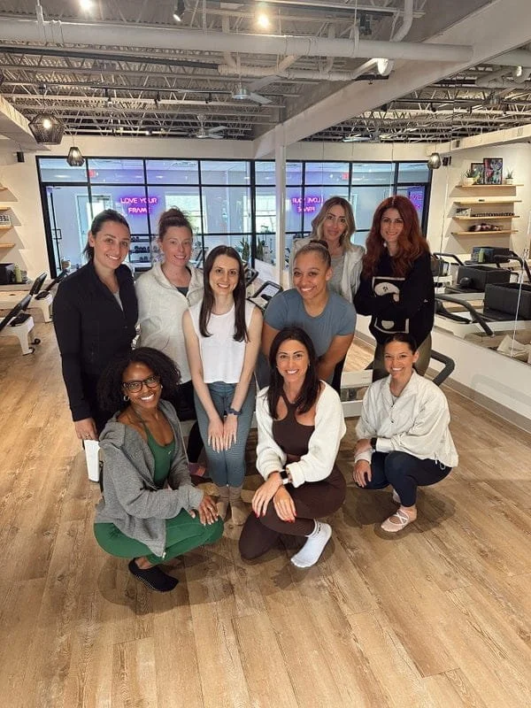 Group of nine women posing inside a modern gym or fitness studio with exercise equipment in the background.
