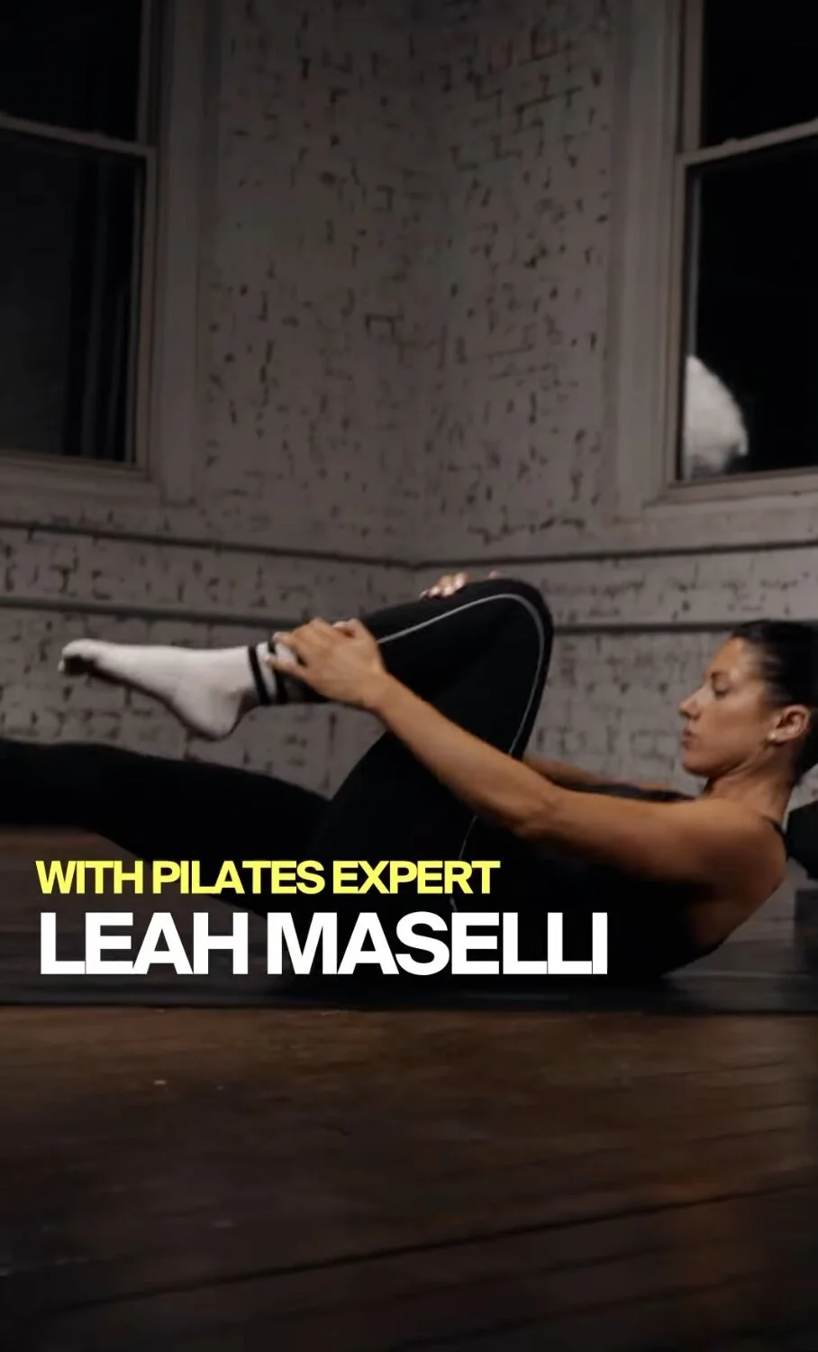 A woman in workout clothes performs a pilates exercise on a mat in a studio with white brick walls and windows at night. Text overlay: "WITH PILATES EXPERT LEAH MASELLI."