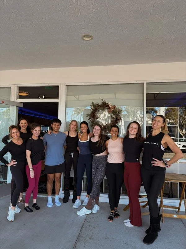 Group of ten women and one man standing outside in front of a glass door with a holiday wreath, smiling, posing for a photo.