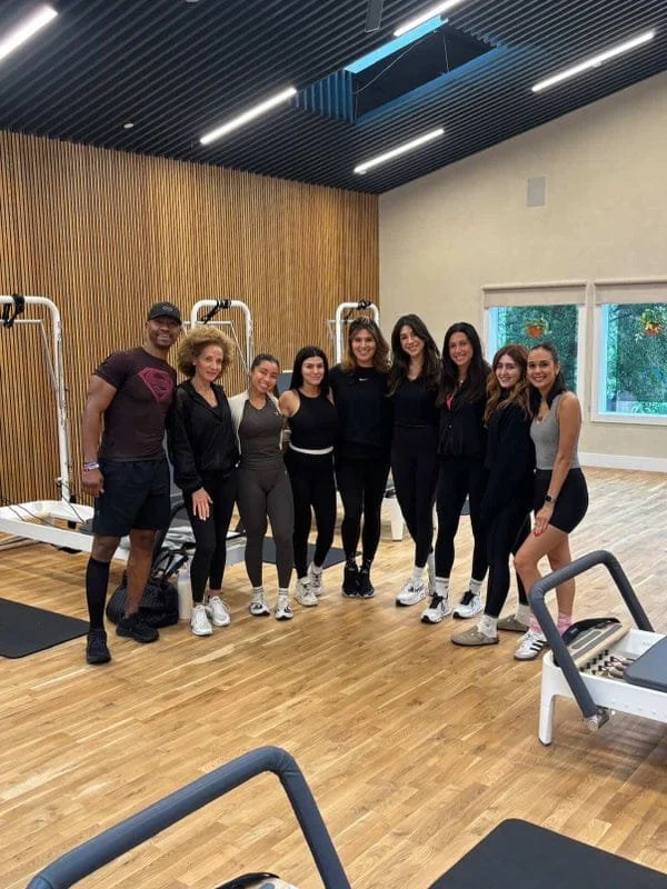 Group of eight women and one man standing together in a gym, smiling for the photo. There are exercise machines and a wooden wall in the background, and windows showing trees outside.