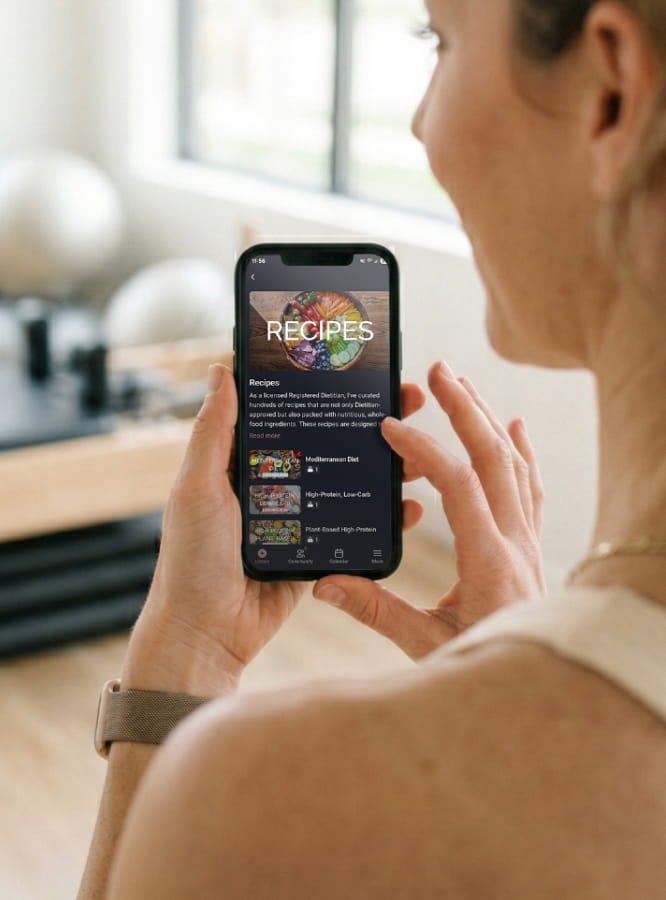 A woman holding a smartphone showing a recipes app or website with categories including Mediterranean diet, high-protein, low-carb, and plant-based options, in a bright kitchen with large windows.
