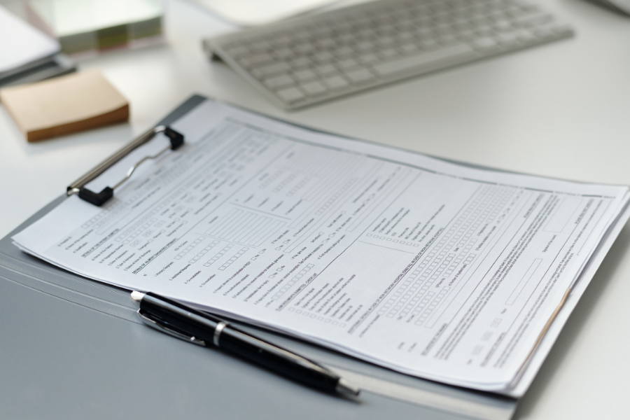 A desk with a clipboard holding a printed form, a black pen, a computer keyboard, and sticky notes.