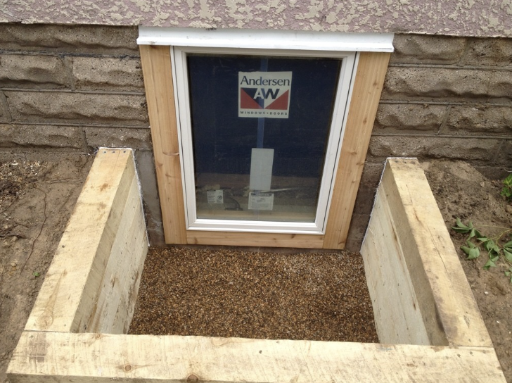 A new egress basement window installed in a brick wall, with a wooden frame and a gravel bed in front, surrounded by garden soil.
