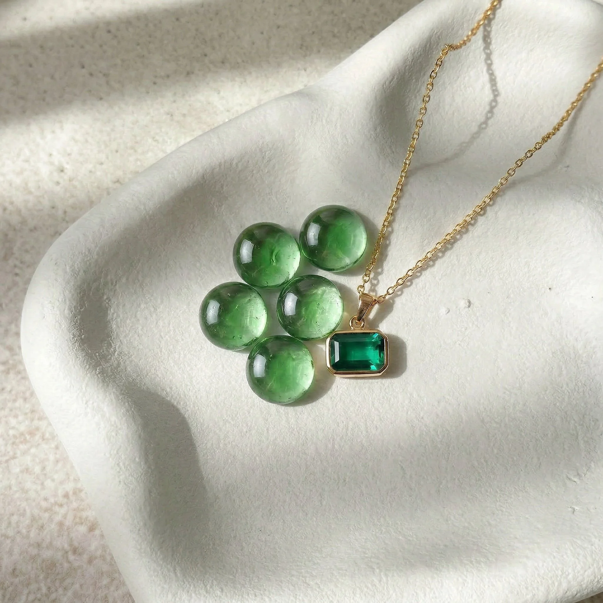 Several green Selah Stones next to a bespoke gold necklace with an emerald-cut green memorial stone pendant on a white ceramic plate.