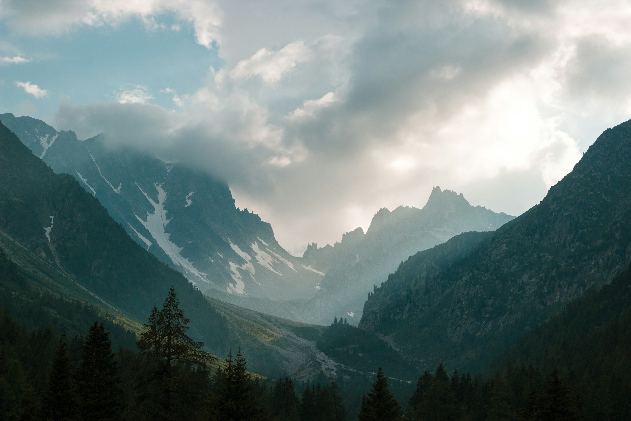 A mountain valley with peaks covered in snow, lush green trees in the foreground, and a partly cloudy sky with sunlight breaking through clouds.