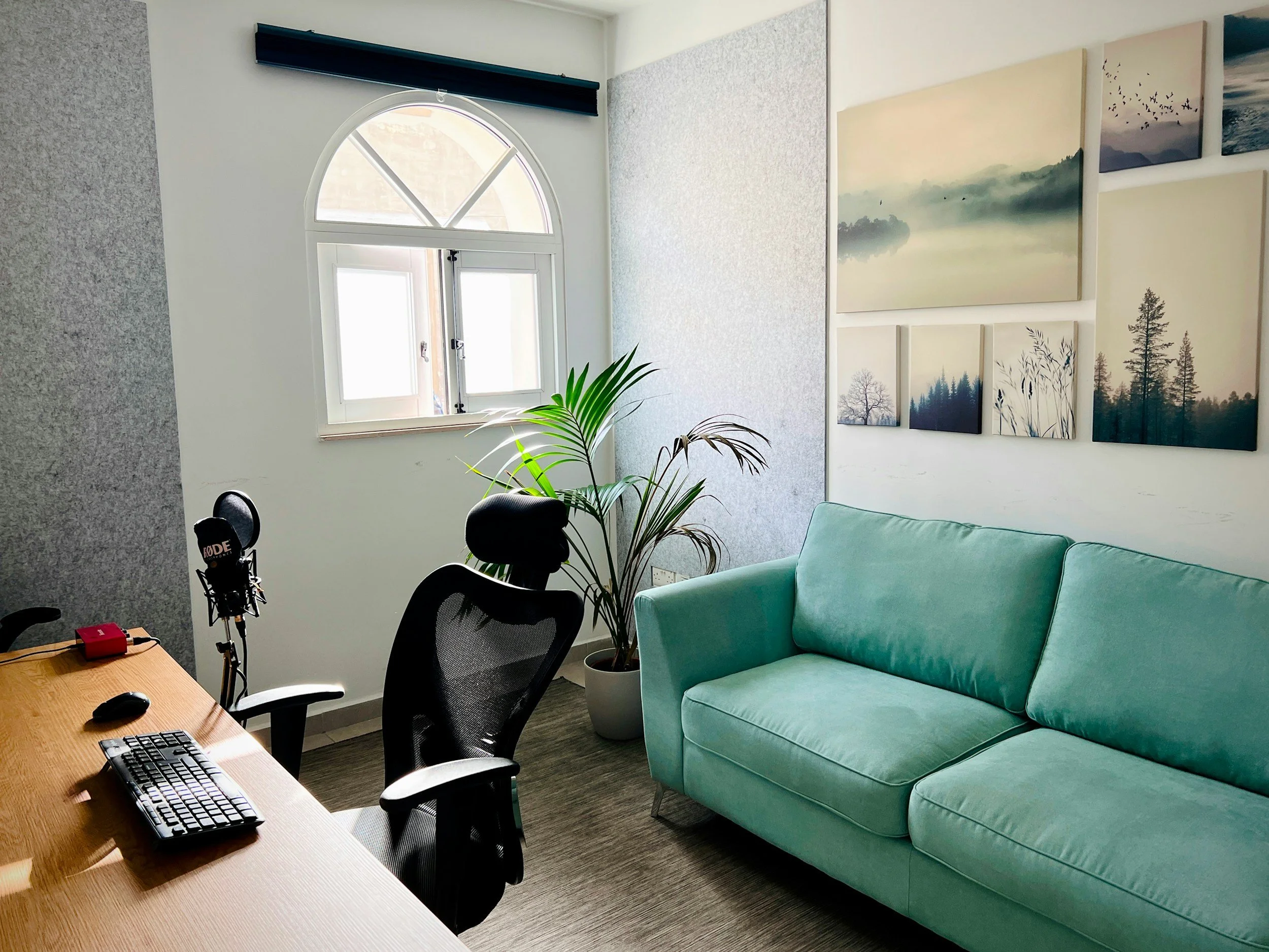 Office space with a wooden desk, black mesh chairs, a computer keyboard, and a microphone. There is a curly plant in a white pot and a mint green sofa. The wall features a gallery of landscape paintings, a window with a semi-circle top and rectangular bottom, and a textured wall panel.
