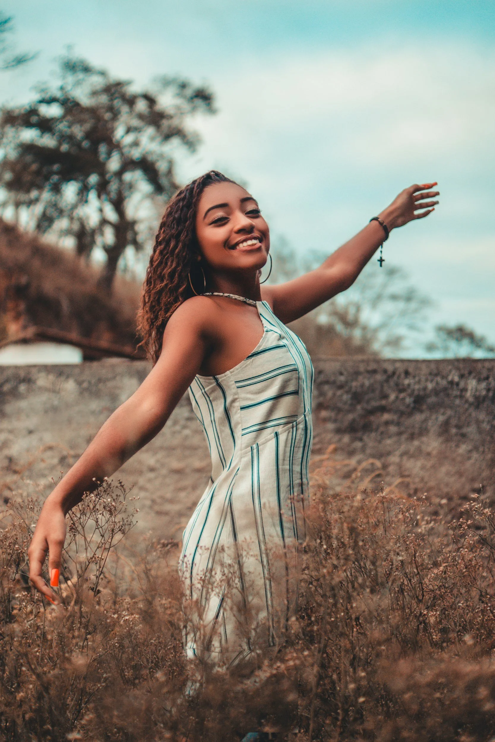 Young woman with curly hair smiling and extending her arm, wearing a striped dress, outdoors in a natural setting with trees and dry grass, during the day.