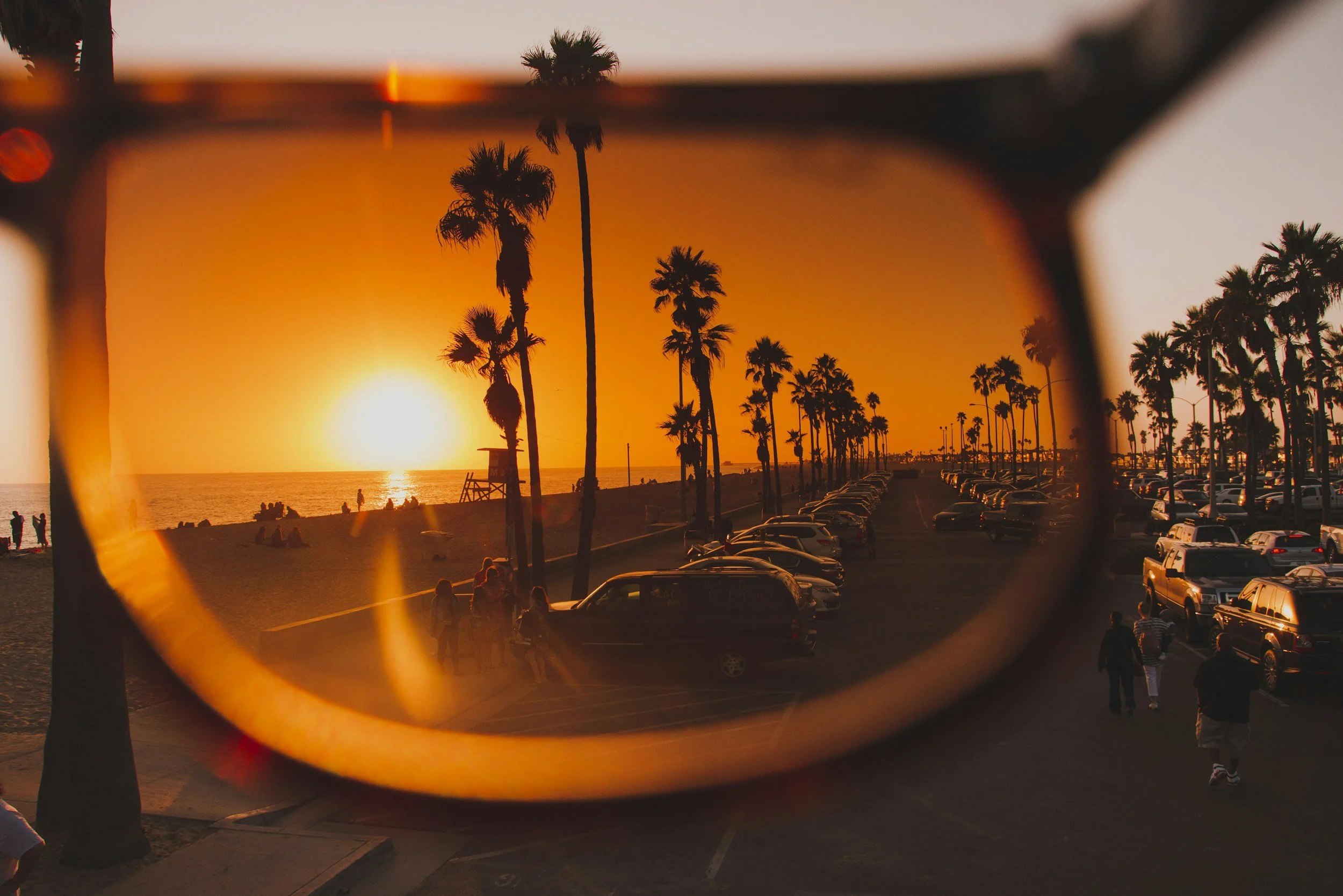 Sunset over a beach viewed through sunglasses with tall palm trees and a parking lot filled with cars in the background.