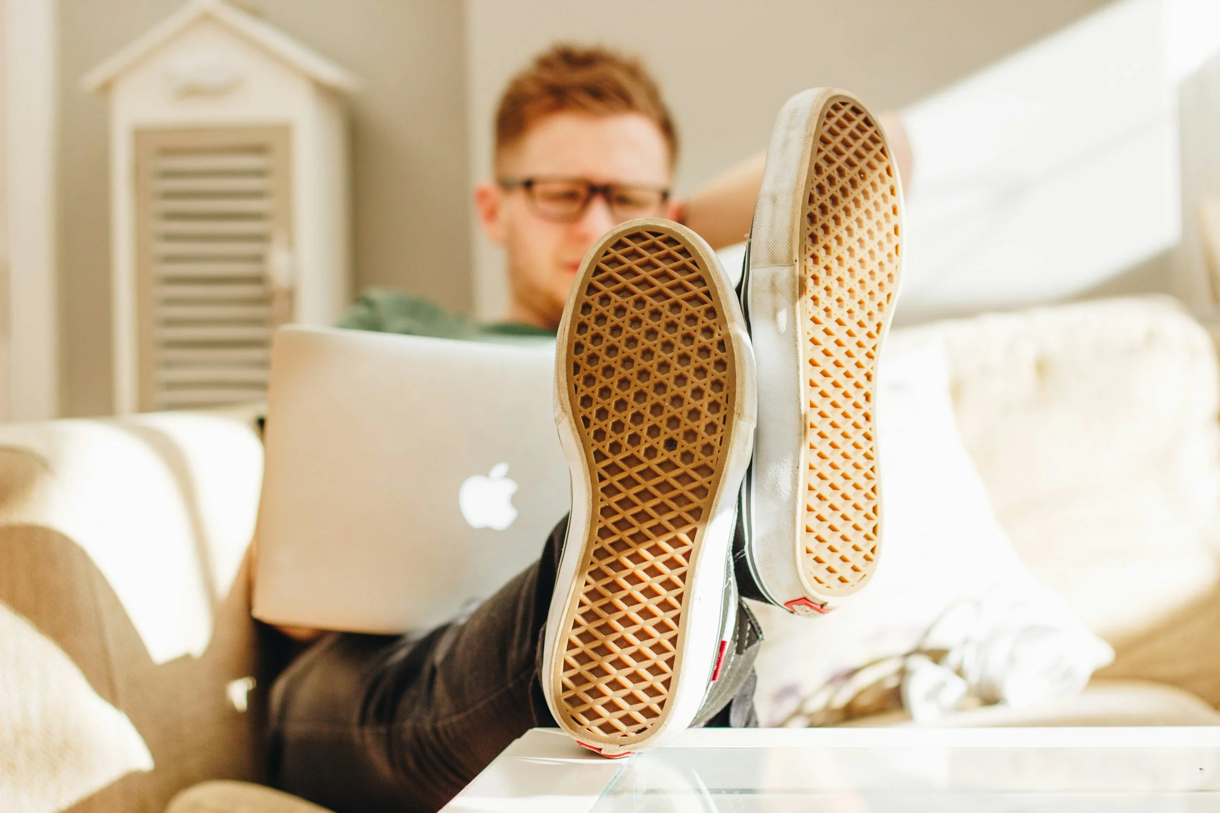 Person with glasses, sitting on a couch, with feet up on a table, wearing sneakers, using a laptop, in a bright room.