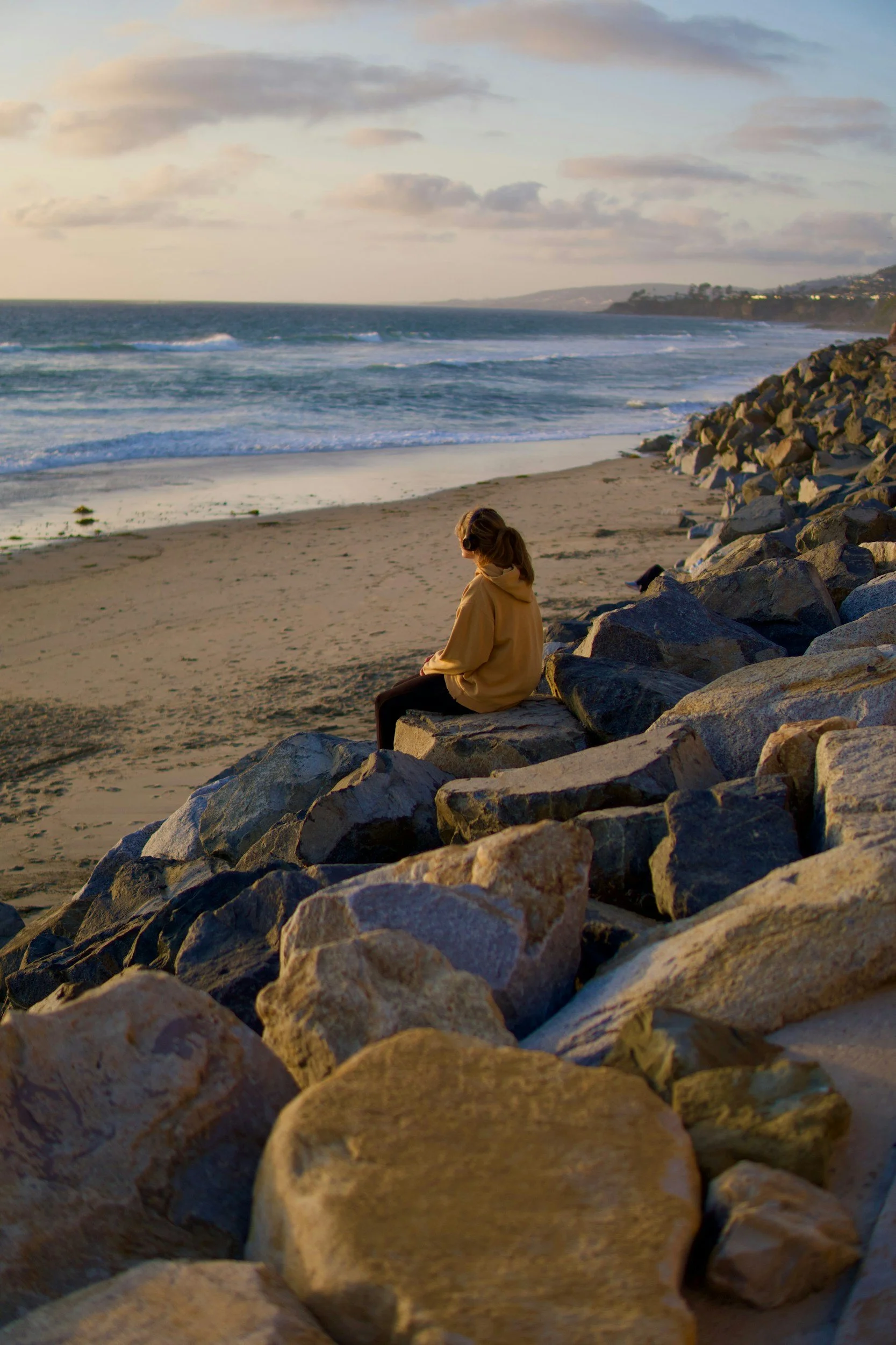 A woman sitting on rocks at the beach, looking out at the ocean during sunset with clouds in the sky.