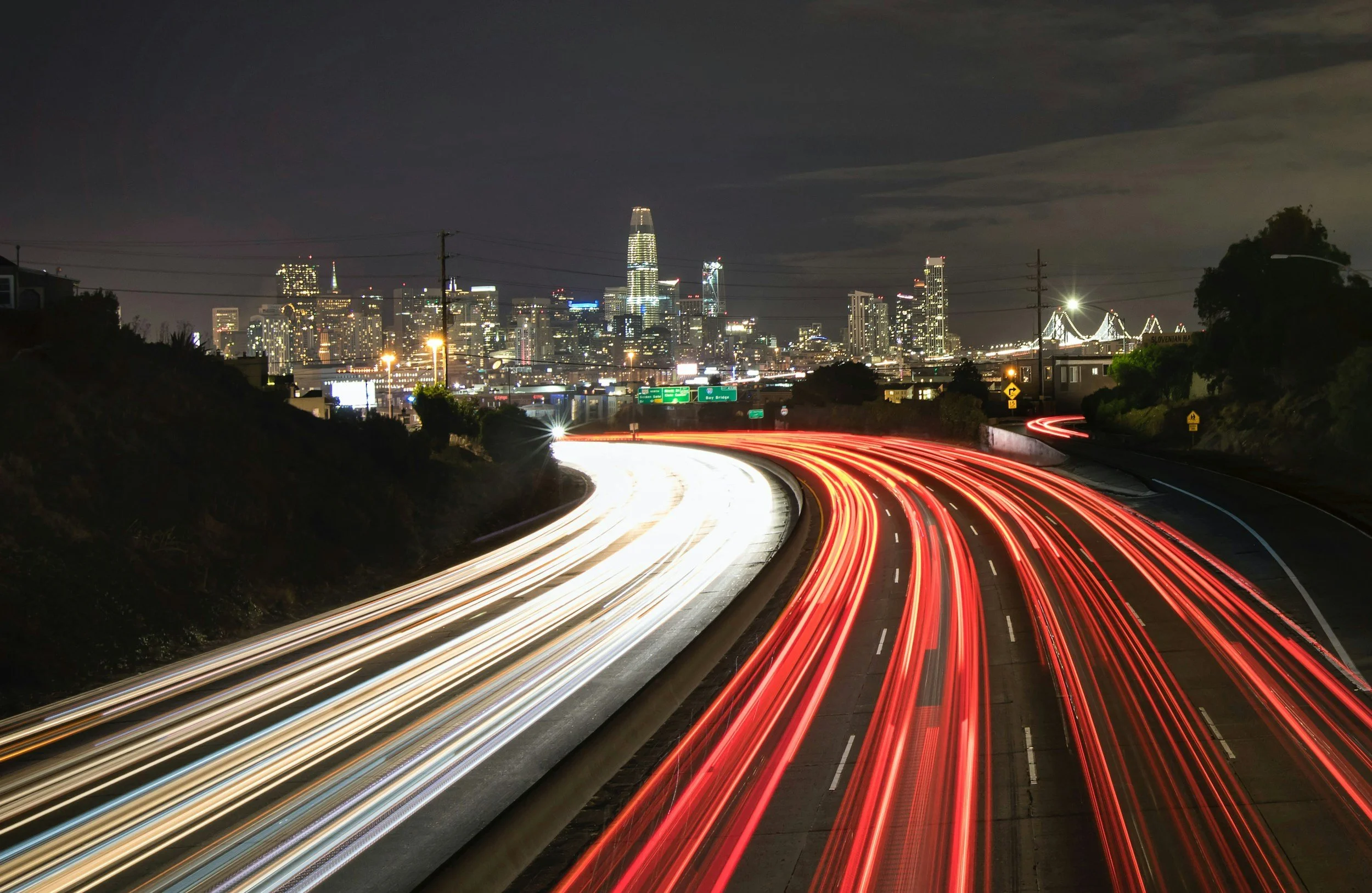 Nighttime view of a city skyline with tall buildings illuminated, showing light trails from moving vehicles on a highway in the foreground.