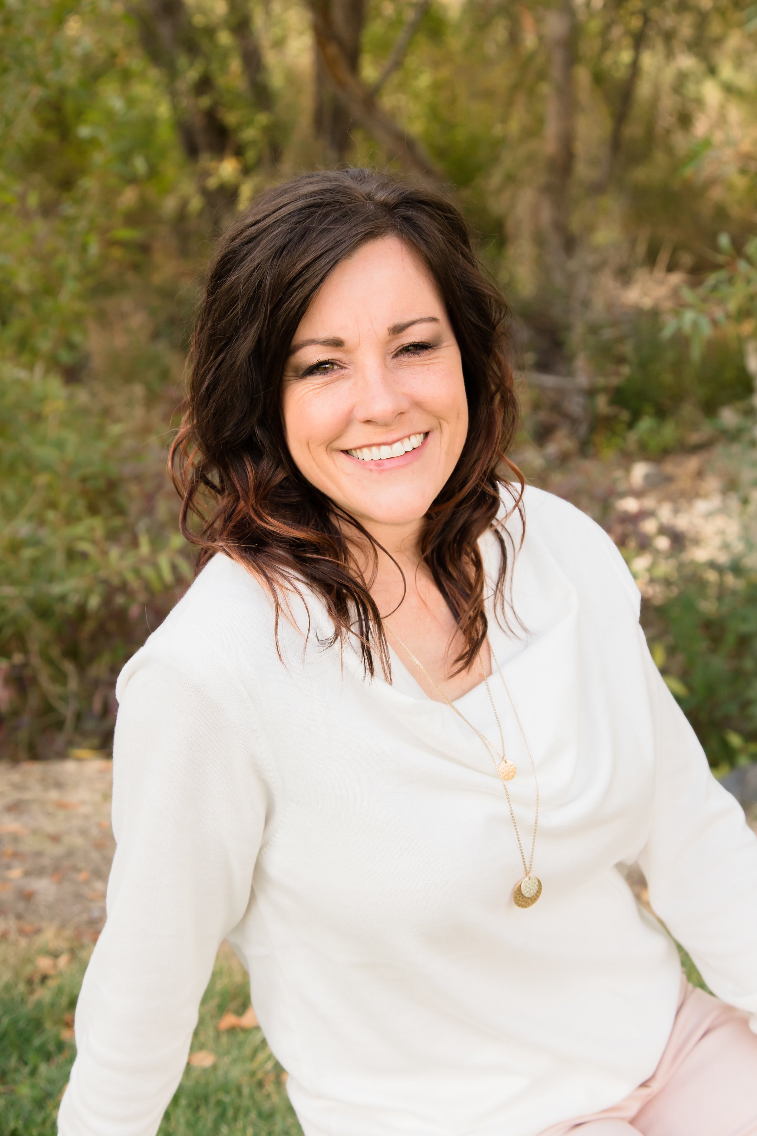 A smiling woman with wavy brown hair sitting outdoors in a natural setting with trees and green foliage in the background.